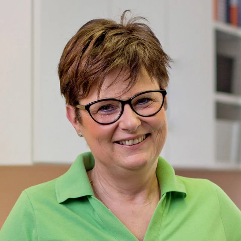 Lächelnde Frau mit Brille und grünem Polo-Shirt im Büro, Hintergrund mit weißen Regalen.