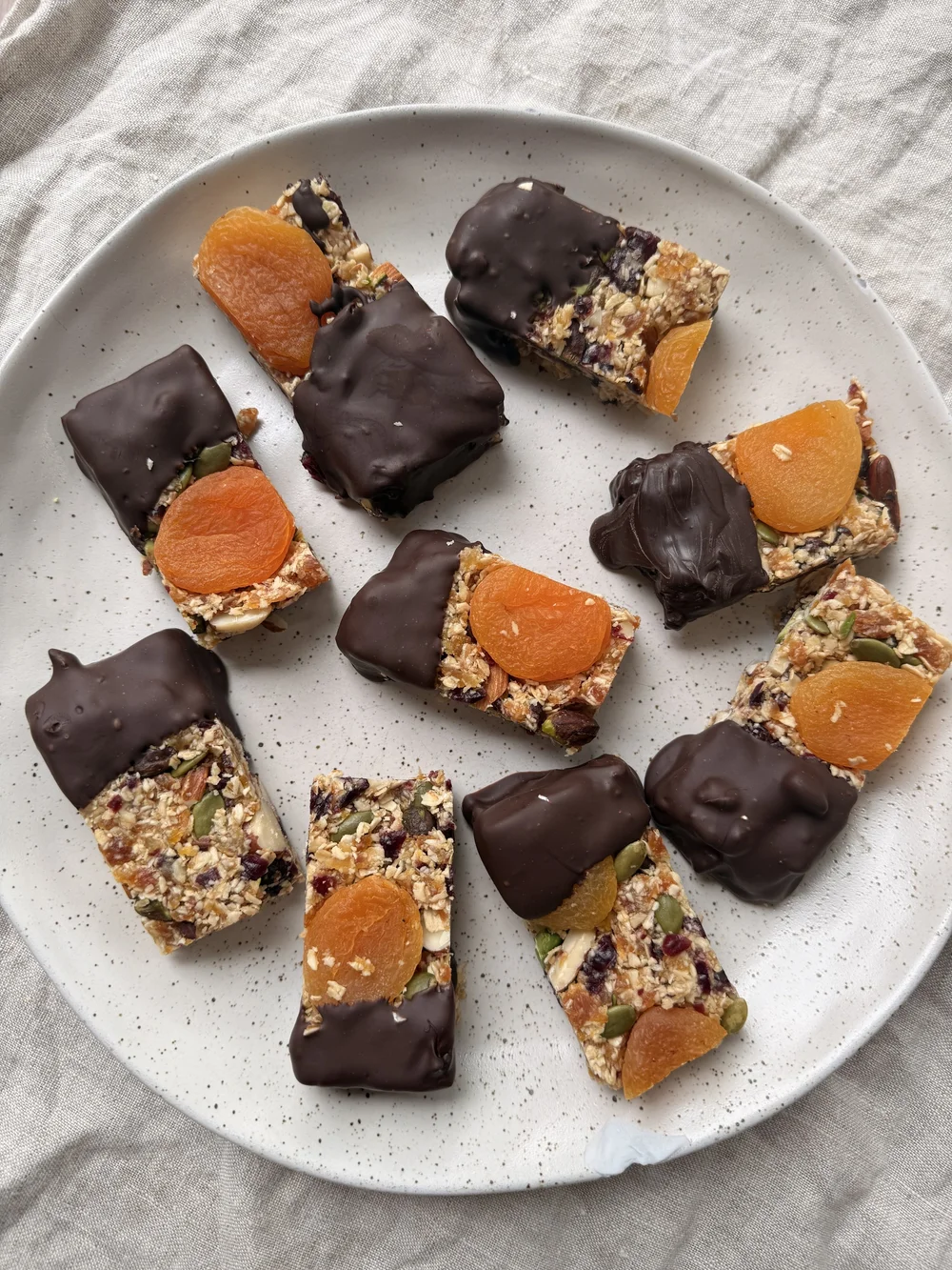Muesli bars on a speckled plate, each topped with a dried apricot and half-dipped in dark chocolate, with oats, pumpkin seeds, almonds and cranberries through them