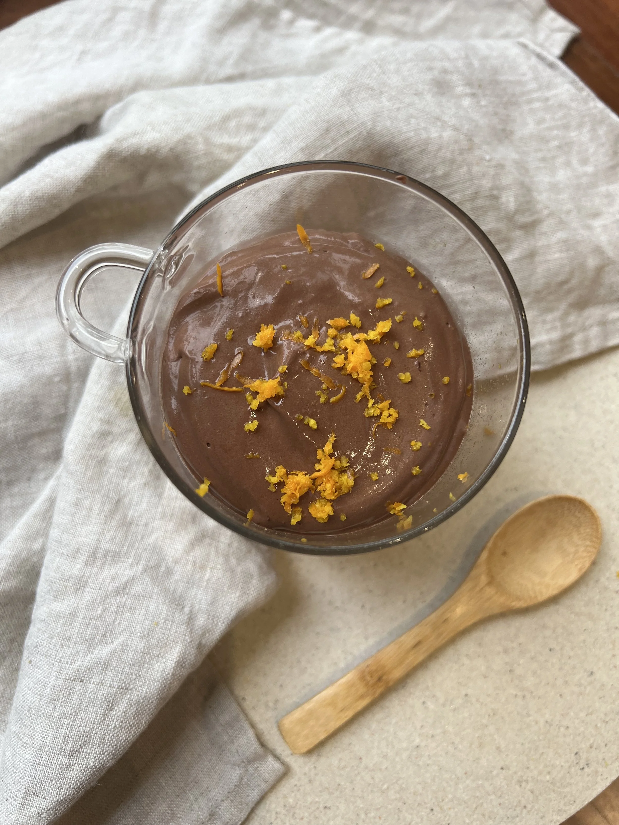 A glass cup of chocolate mousse topped with orange zest, on a beige surface with a wooden spoon and white napkin.