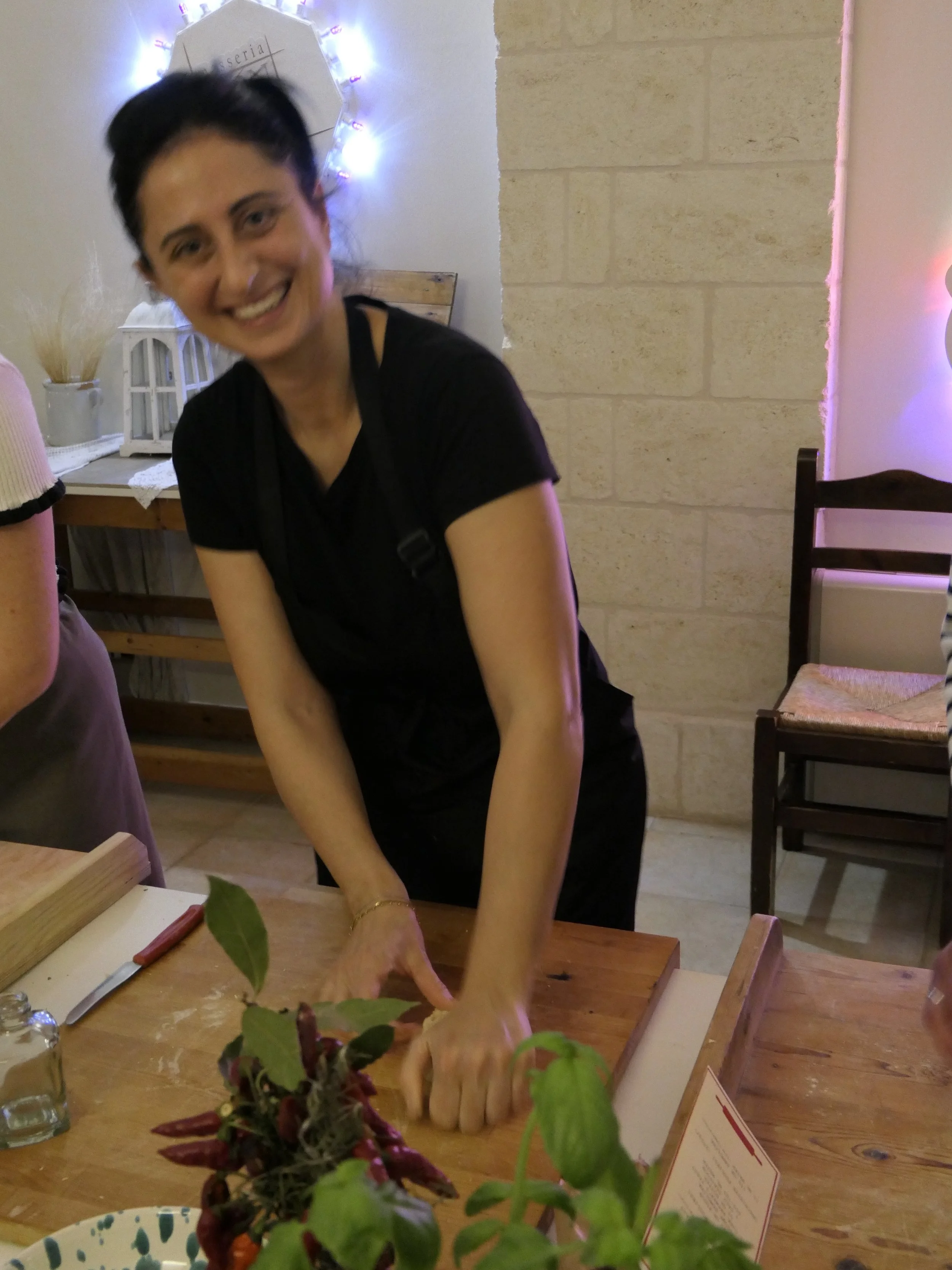 Eine Frau beim Teig ausrollen in einer Küche, lachend, mit dunklem Haar und dunkler Kleidung, umgeben von Küchenutensilien und Pflanzen.