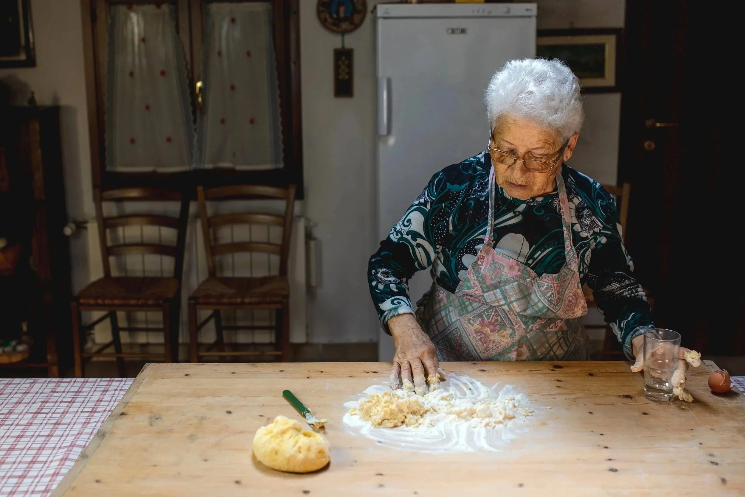 Genussreise-Ältere Frau backt Teig auf einem Holztisch, sie knetet mit der Hand, daneben ein Teigball, ein Messer, ein Glas Wasser und ein Ei.
