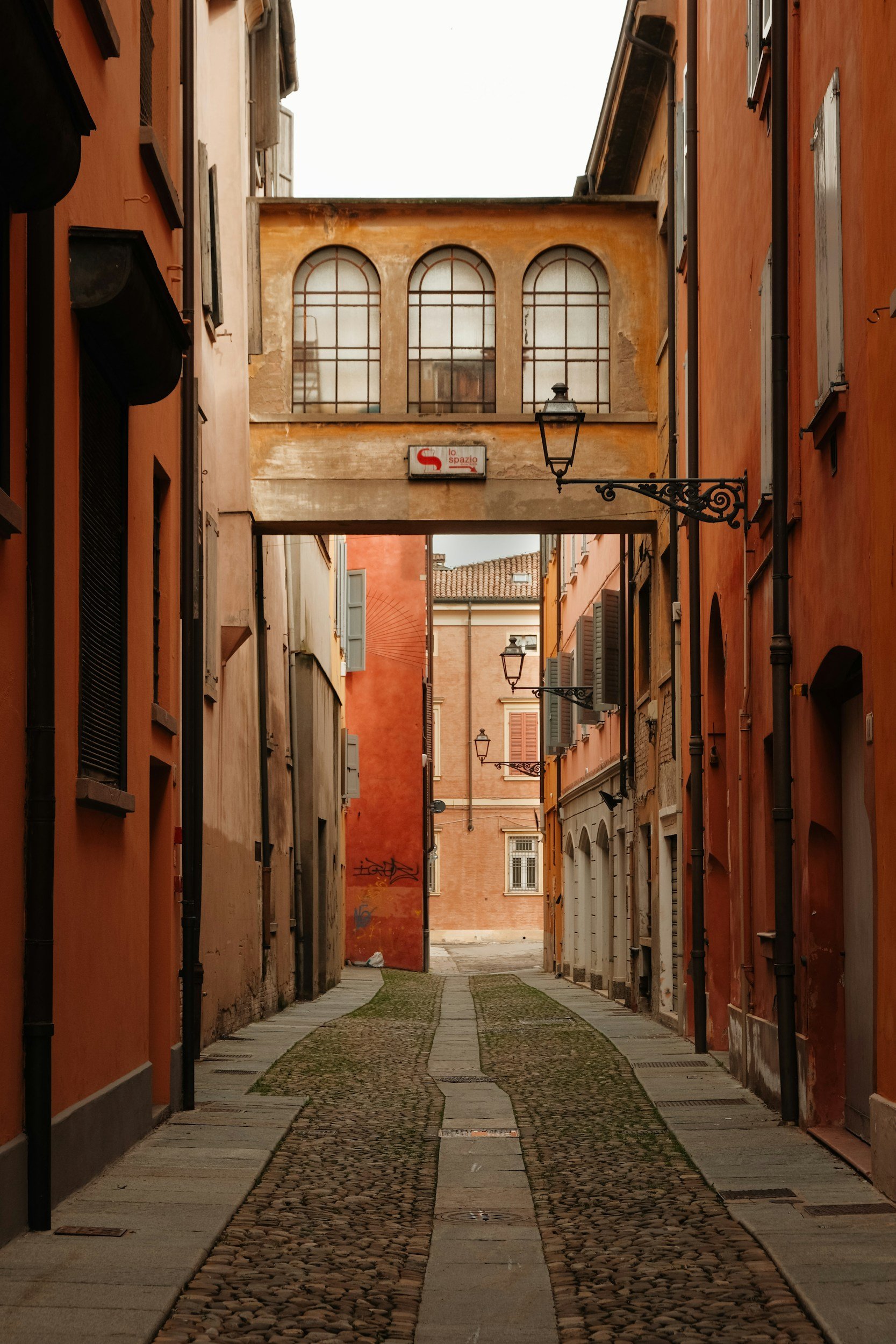 Eine enge, ruhige Gasse mit alten Gebäuden und Straßenlaternen, verbunden durch einen Durchgang mit schrägen Fenstern, in einer italienischen Stadt.