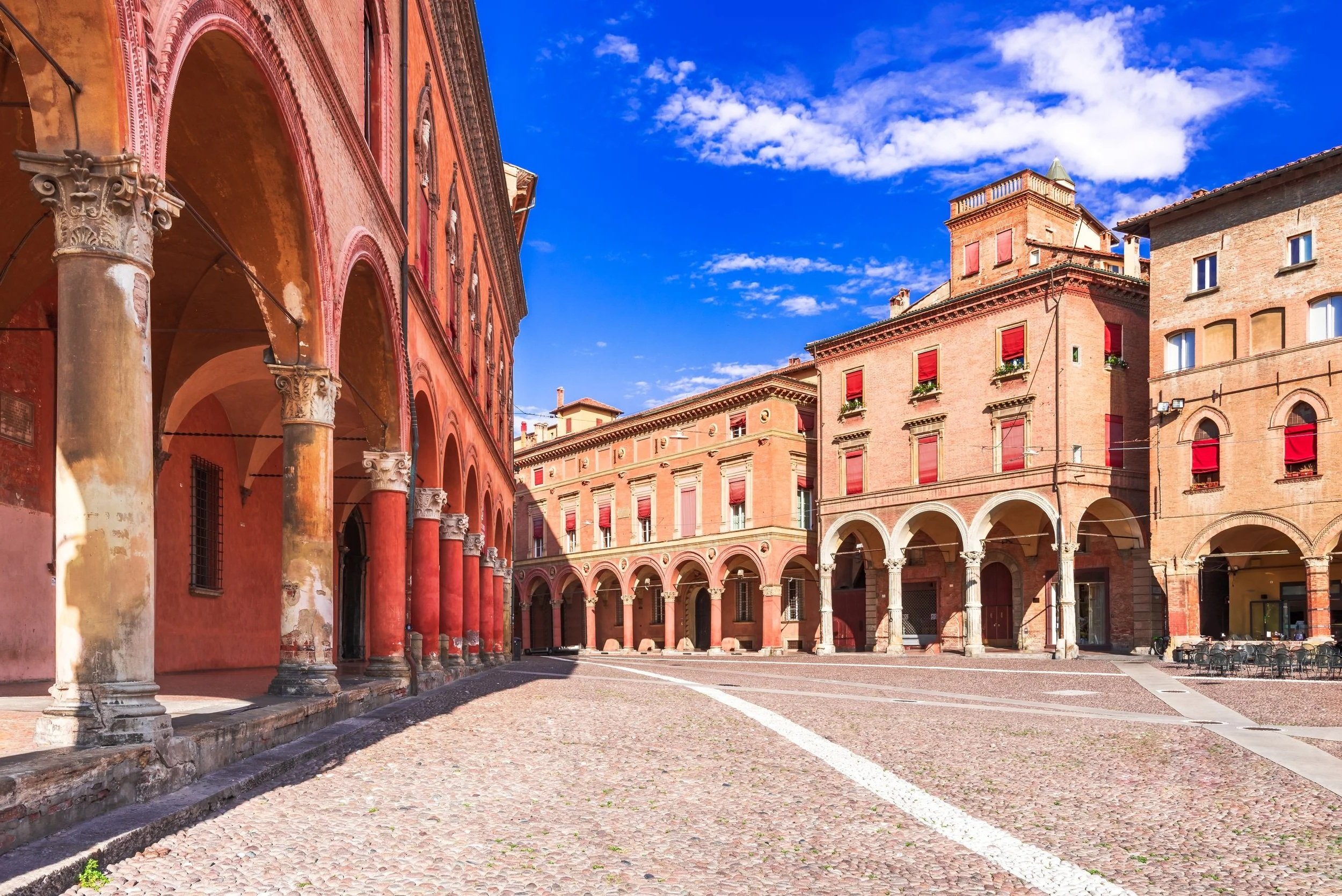 Historische Stadt Bologna mit roten Gebäuden und Arkaden, gepflasterter Platz unter blauem Himmel.