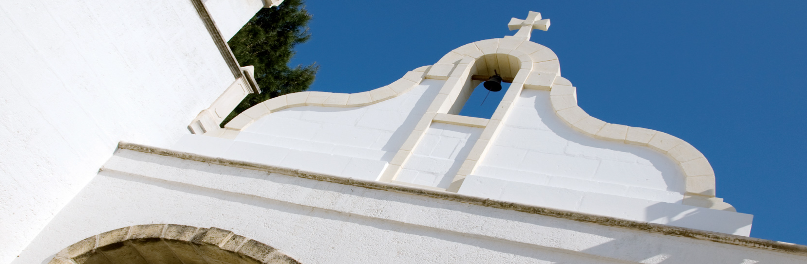 Weiße Kirchenfassade mit Glockenturm und Kreuz gegen blauen Himmel.