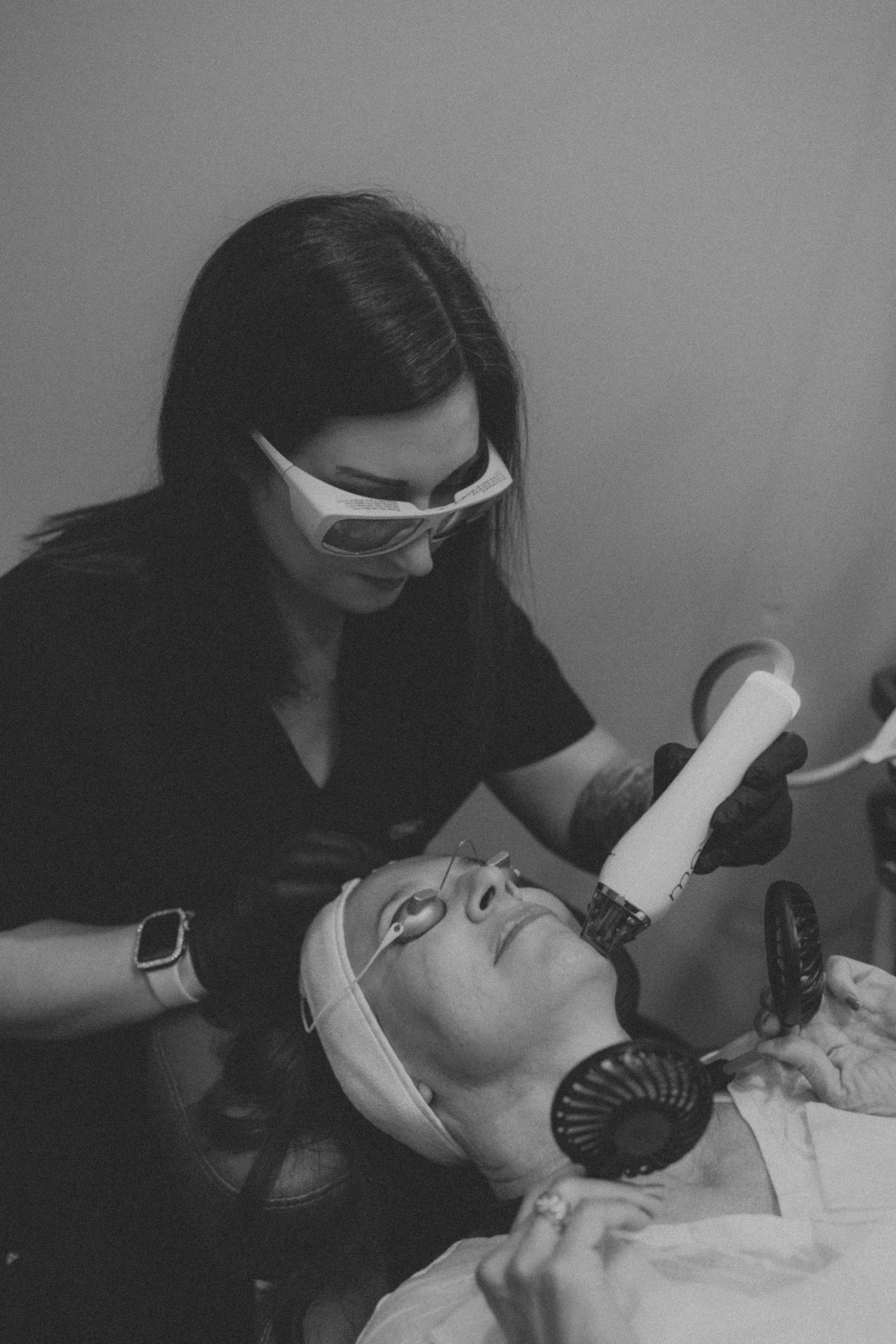 A healthcare professional with dark hair and sunglasses performing a cosmetic or medical tattoo procedure on a patient with medical equipment. Both are lying down; the professional holds a tattoo machine, and the patient wears a bandage on her forehe