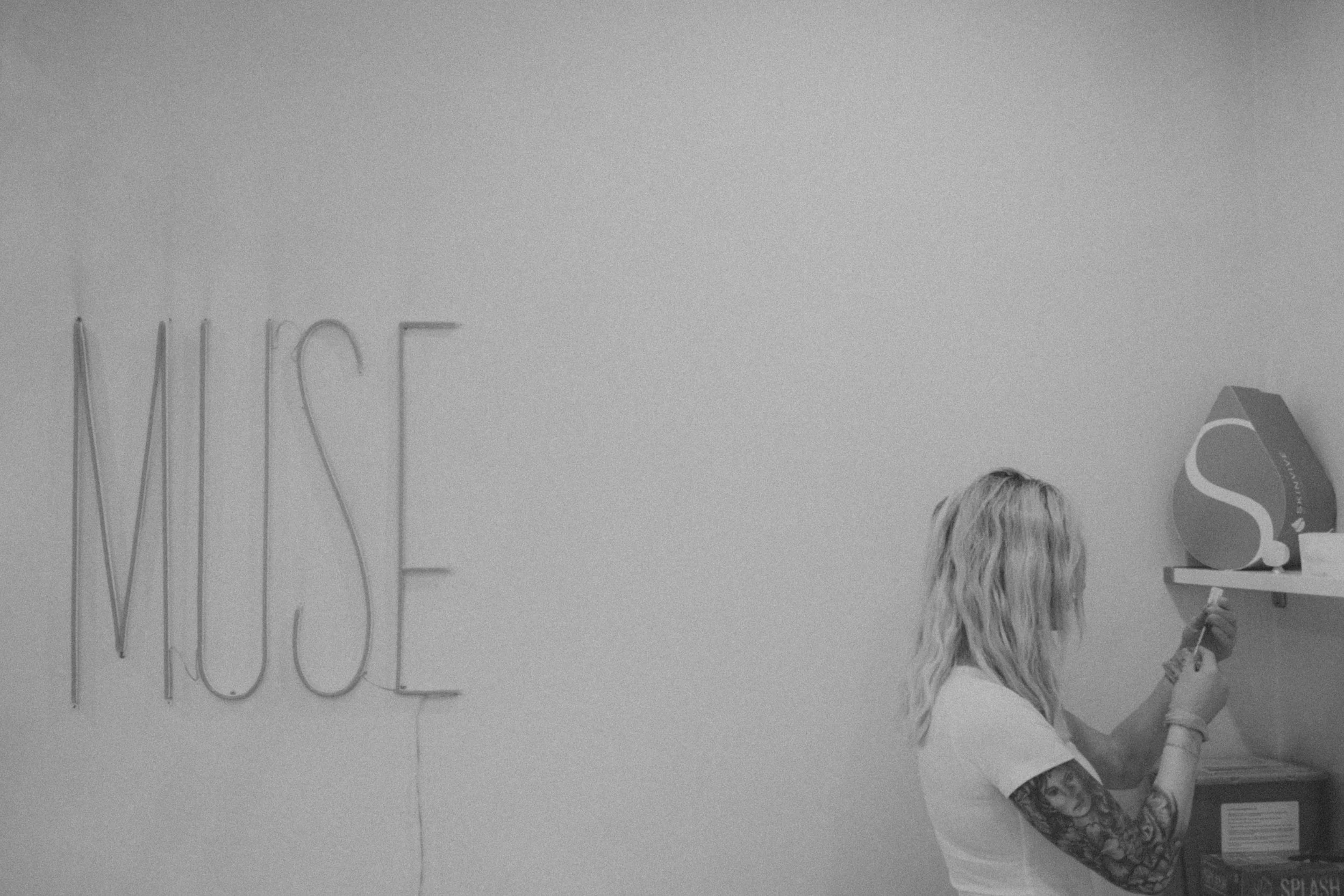 A woman with long curly hair holding an acrylic sign with the text 'SPLASH' on it, standing in front of a wall with the partial word 'NICE' in neon lights, in an indoor setting.