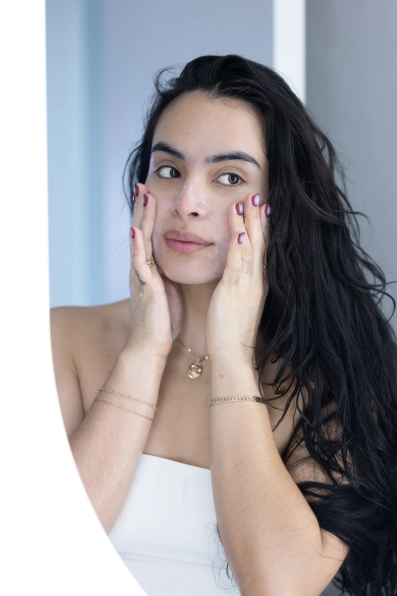 A woman with long, dark, wavy hair is touching her face with both hands, applying a facial product. She has blue eyes and is wearing gold jewelry, including rings, a necklace, and bracelets. The background is a soft, light-colored wall.