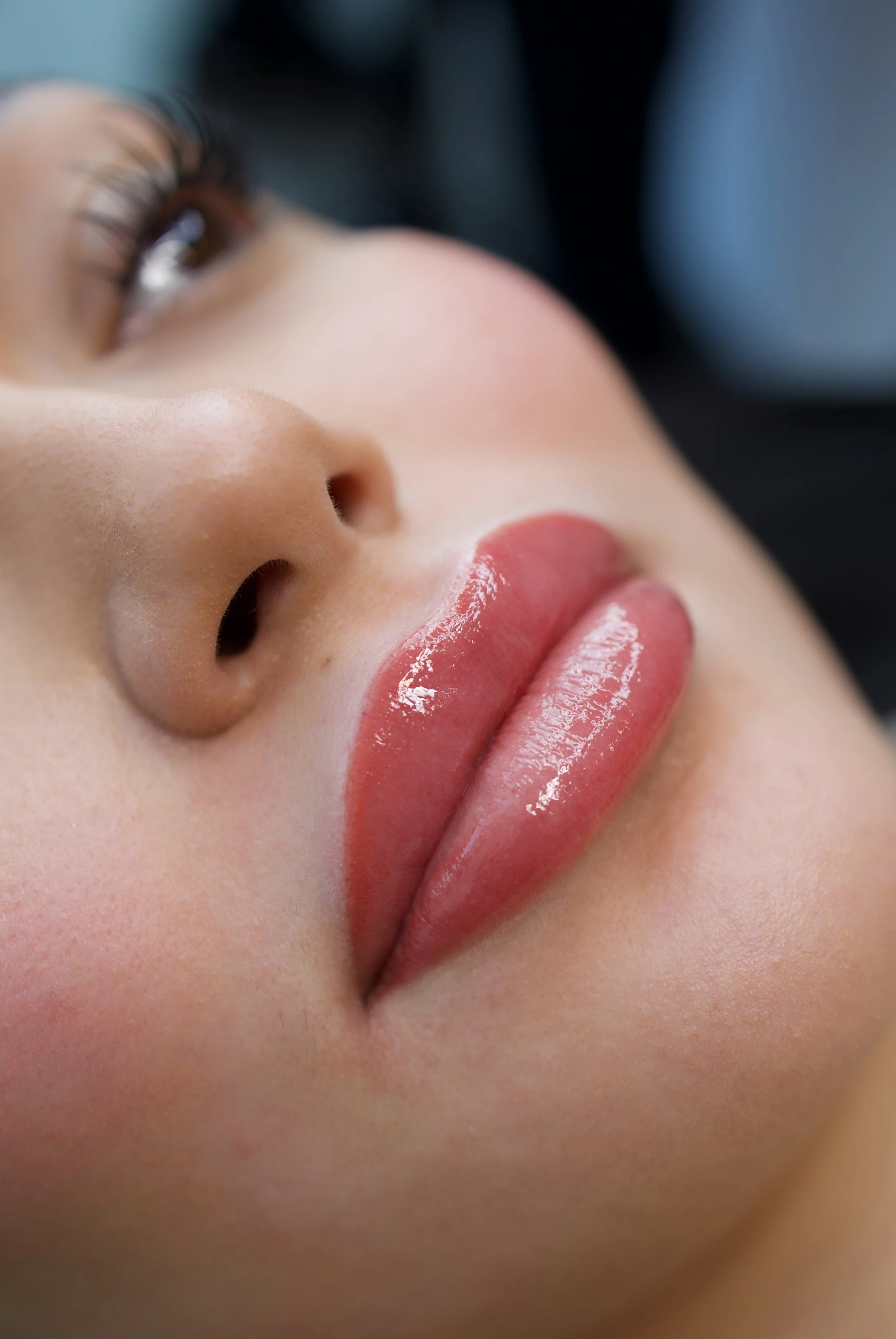 Close-up of a woman's face showing her eye, nose, and lips; her lips are slightly parted and glossy, with her tongue touching her upper lip.