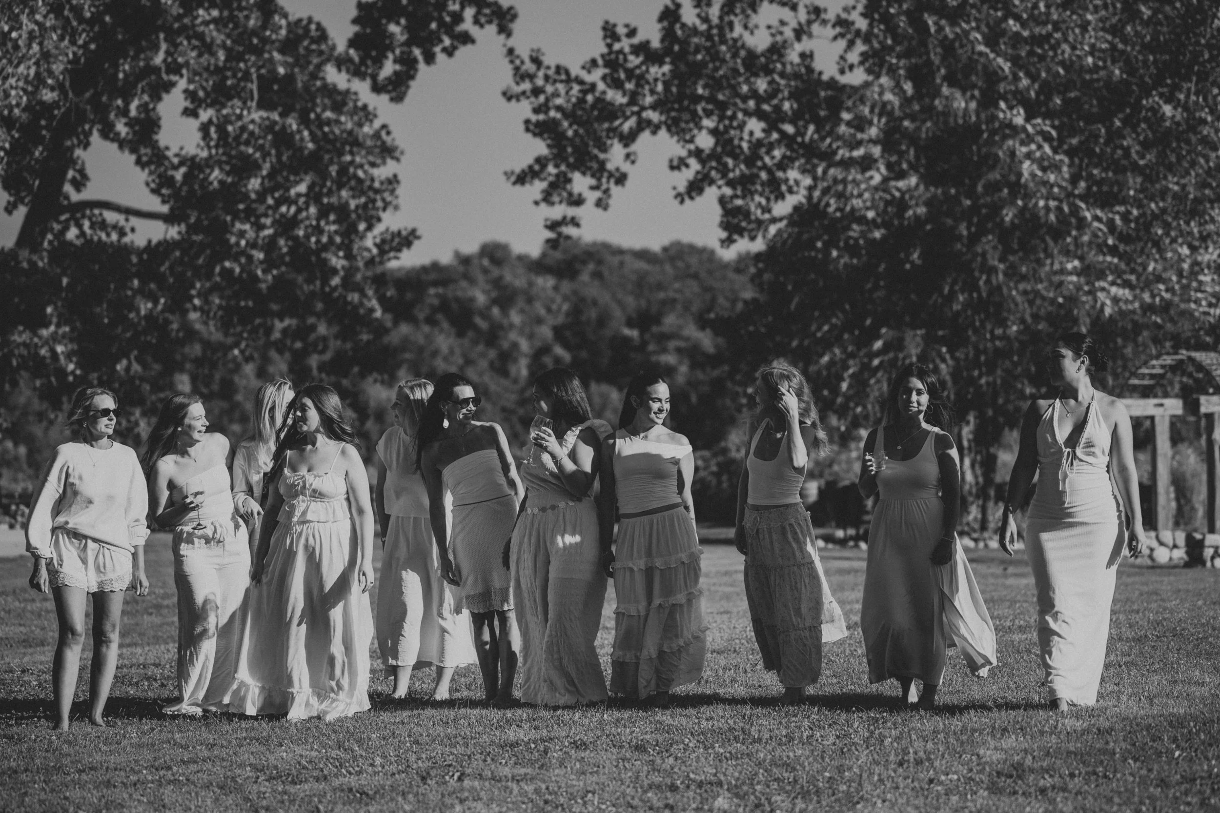 Black and white photo of a group of women in summer dresses walking and talking outdoors in a park with trees in the background.