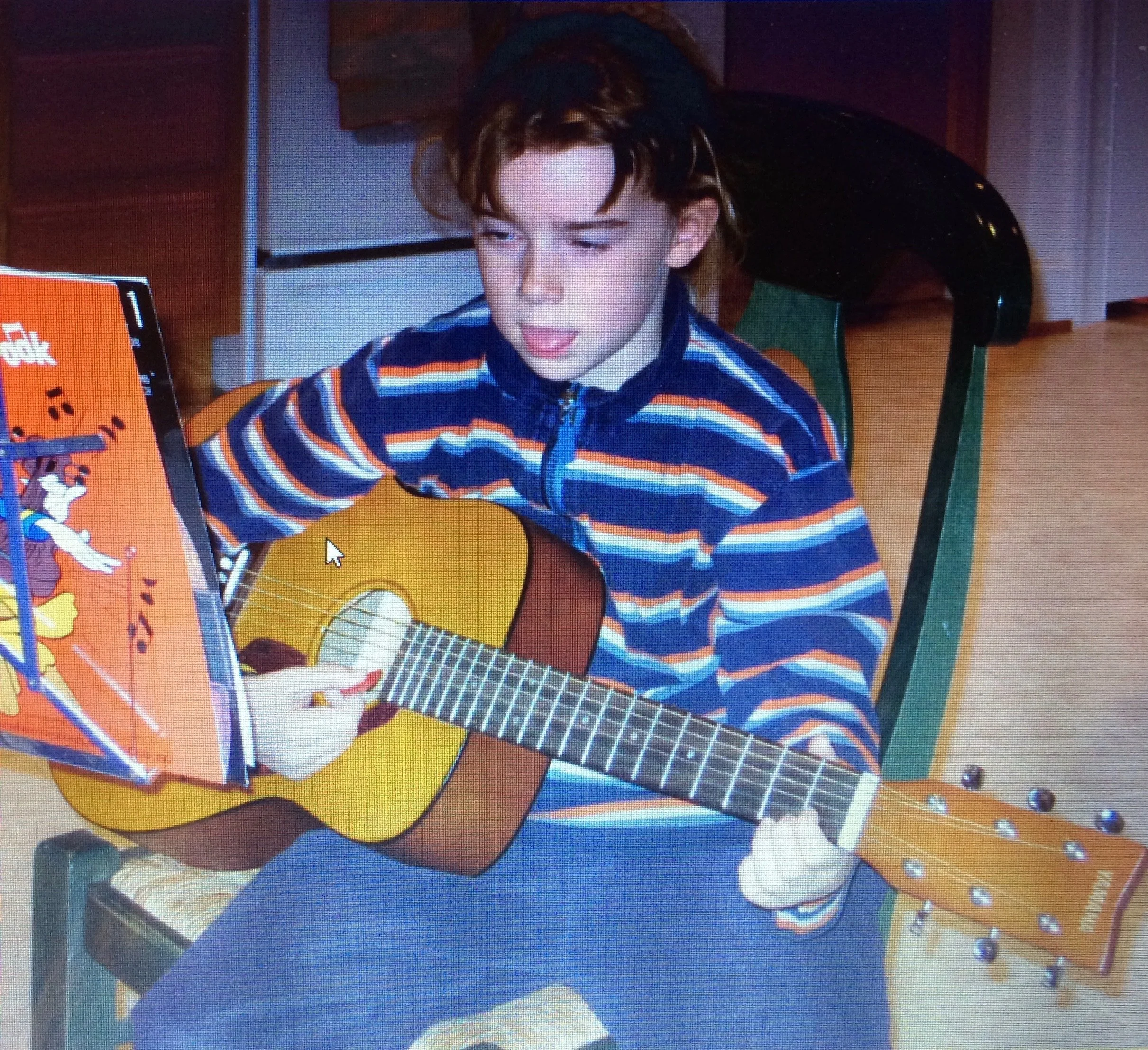 A young person with wavy brown hair around shoulder-length, wearing a striped zip-up jacket, playing a yellow acoustic guitar.