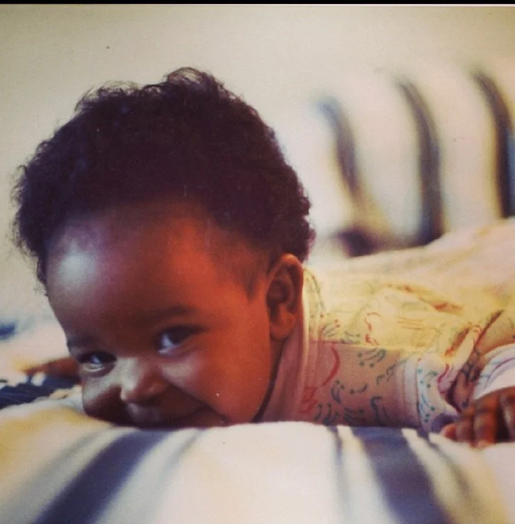 A young child with dark curly hair lying on a bed, smiling and looking at the camera.