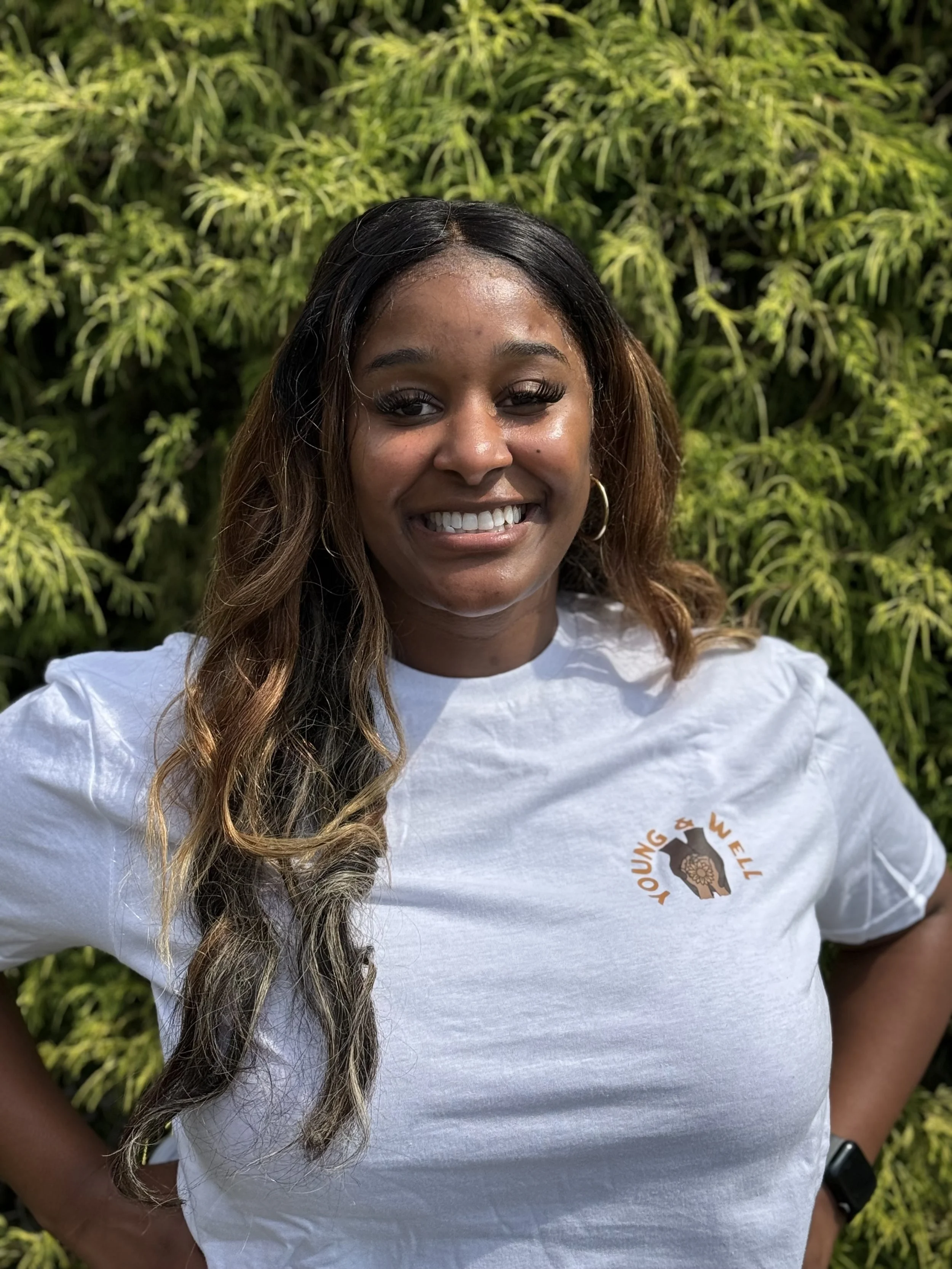 A young woman smiling outdoors in front of green plants, wearing a white t-shirt with a logo and gold hoop earrings.