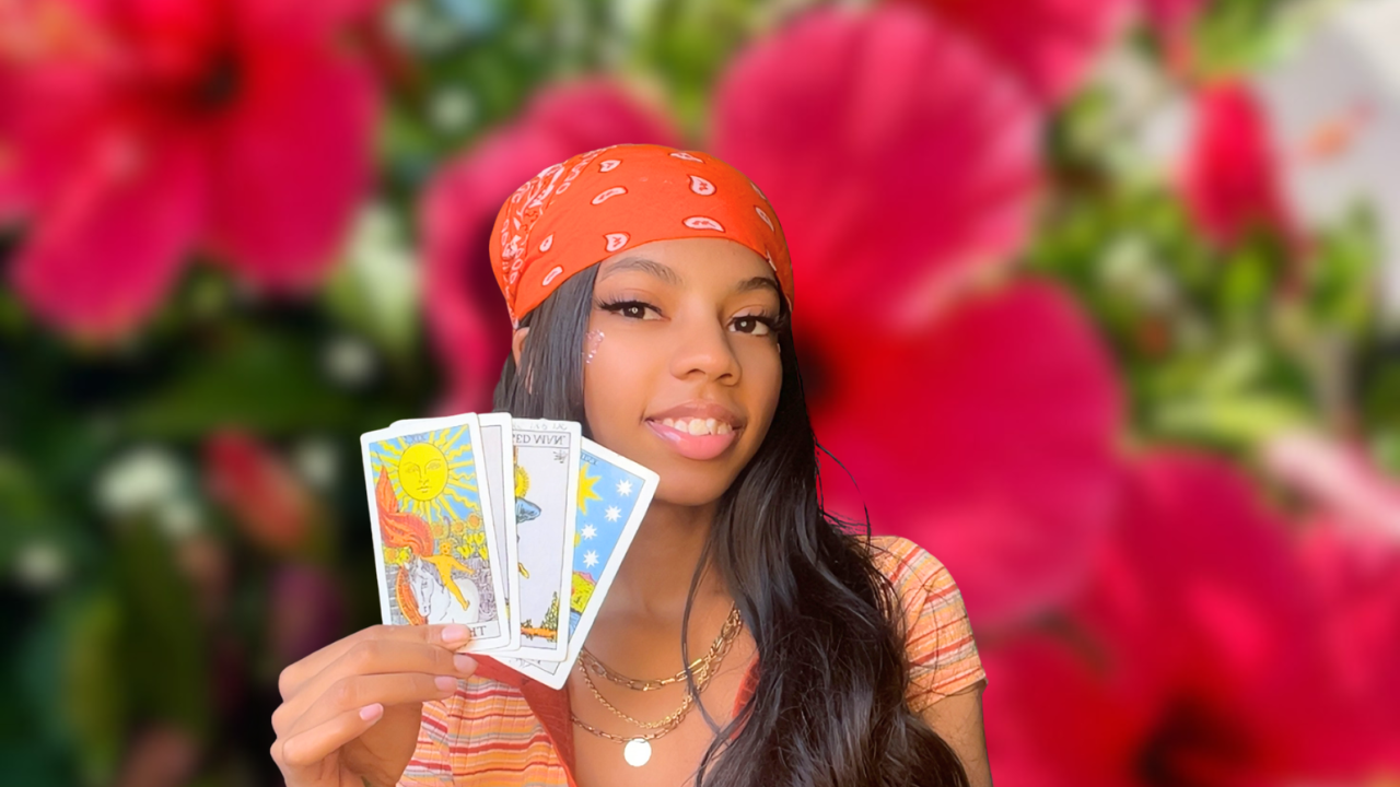 A young woman with long dark hair wearing an orange bandana and a striped top, holding tarot cards, standing in front of a blurred background of pink flowers.