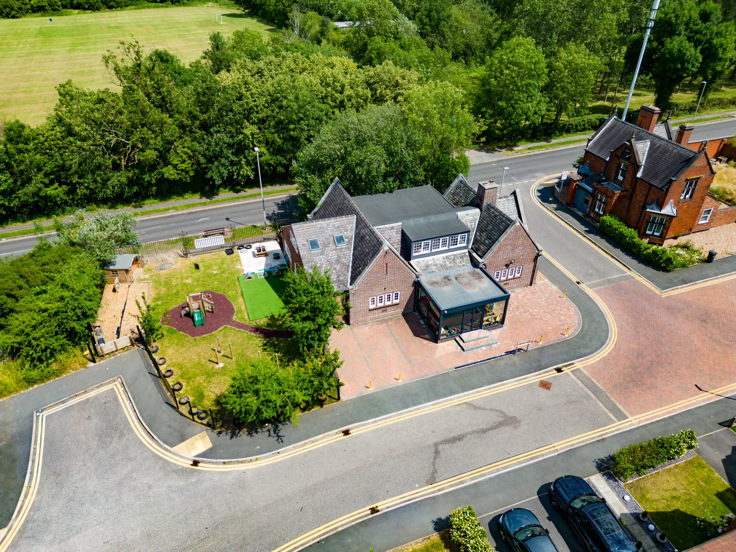 An aerial view of a residential neighborhood with a large house, a small building, and a backyard with a playground, beside a wooded area and a football field in the distance. The streets are paved with yellow and white lines, and there are a few par