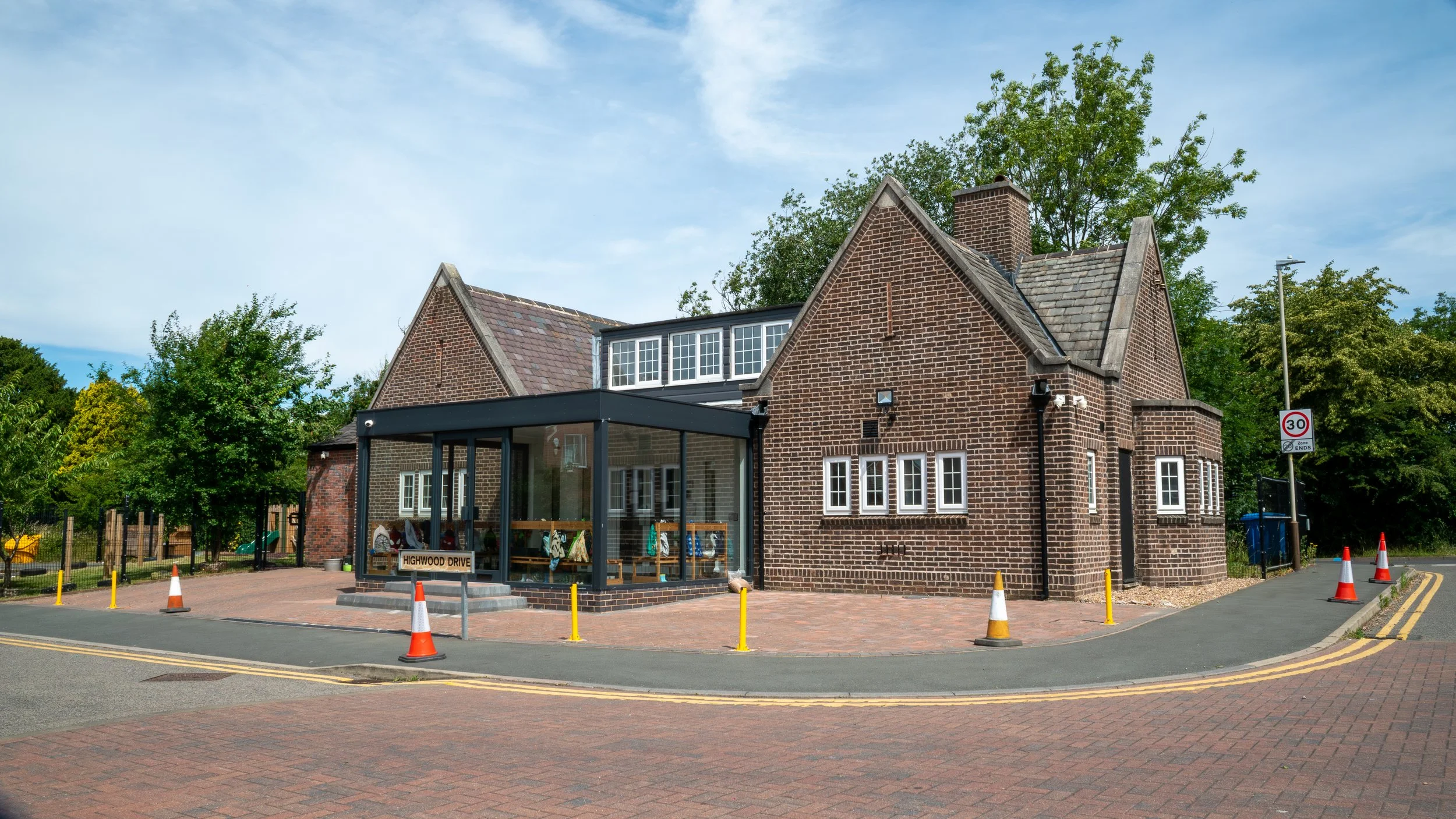 A brick building with a black extension and large windows, surrounded by trees, with a small paved area and traffic cones in front, under a partly cloudy sky.