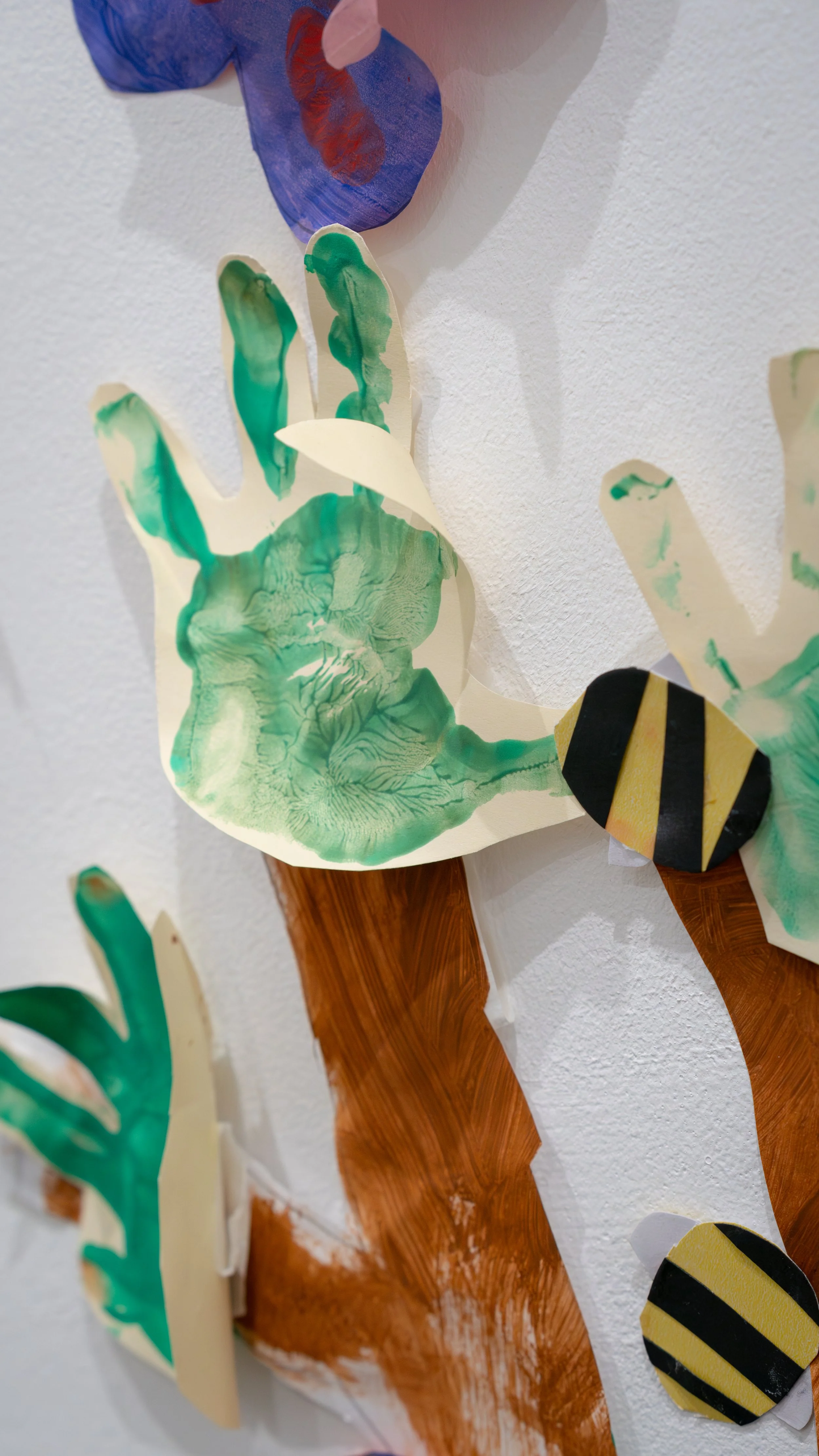 Colorful paper art of a green and black striped bumblebee on a tree branch with green leaves, mounted on a white wall.