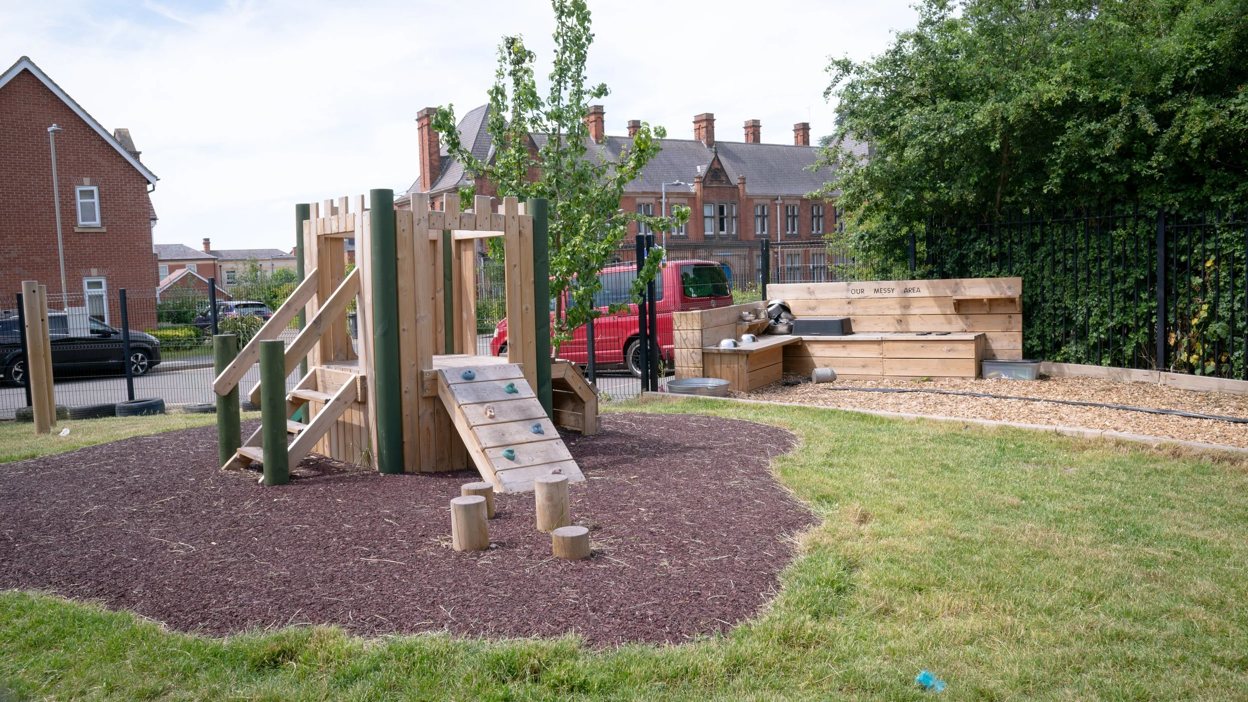 A playground with a wooden climbing structure and a rock wall, surrounded by mulch, with a green grassy area and trees in the background, and a bench with a sign that reads 'Our Messy Area'.