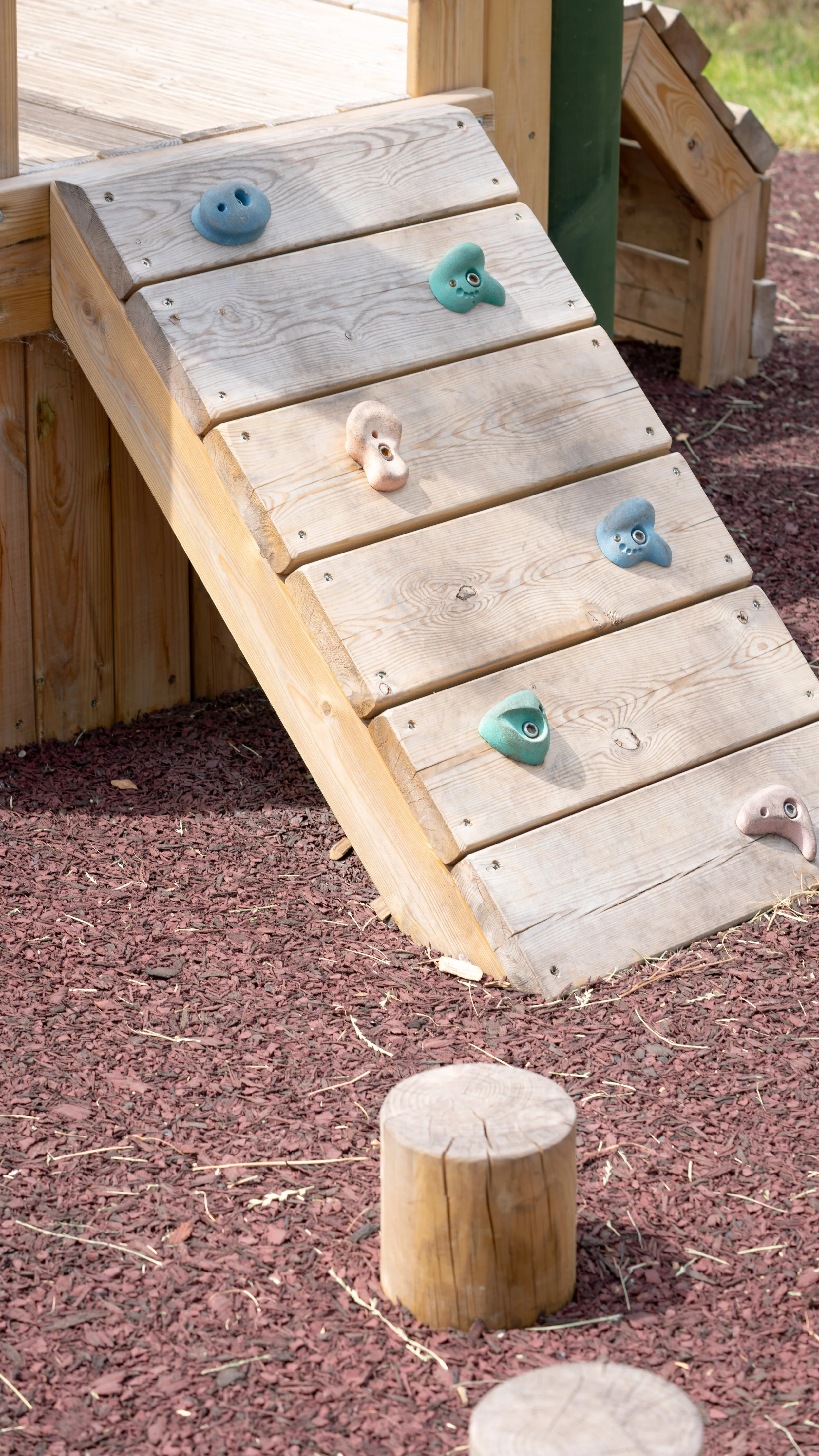 Wooden outdoor climbing wall with colorful handholds, situated on reddish mulch ground, with wooden logs in the foreground.