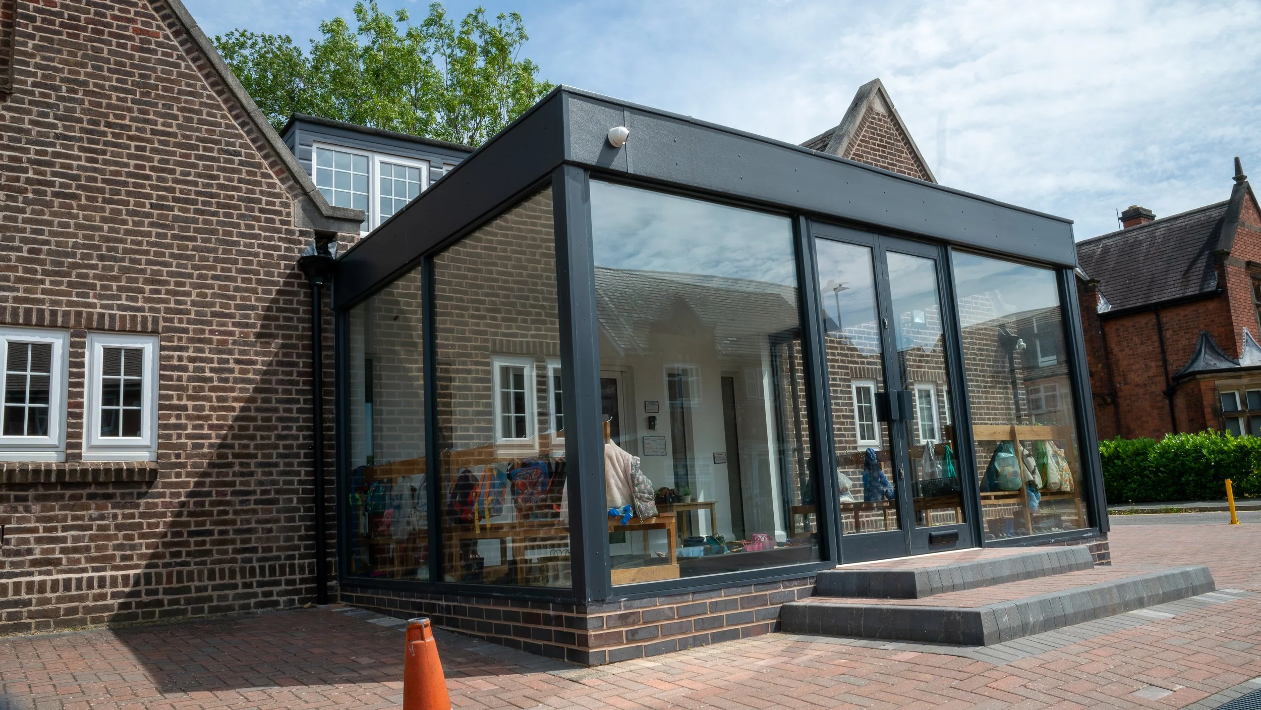 A modern glass and metal extension attached to a traditional brick building, with some interior items visible through the glass, under a partly cloudy sky.
