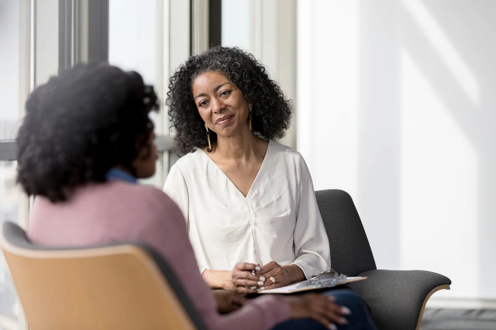 A confidential one-on-one conversation, with two individfuals seated facing each other, one holding a clipboard while listening attentively, suggesting a coaching, counseling, or advisory session.