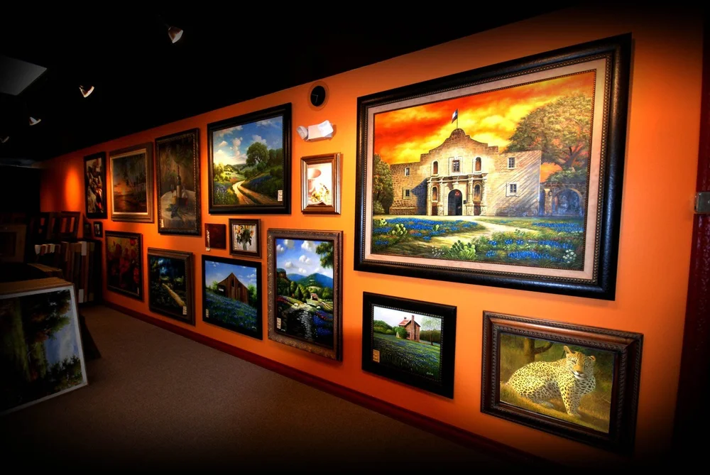 Painting of the Alamo in bluebonnets hanging with Texas Hill Country and wildlife paintings on the orange gallery wall at The Frame Gallery in San Antonio