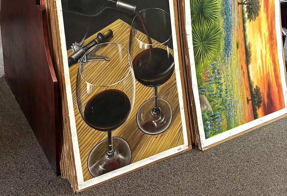 Unframed hand-painted canvases of wine glasses and bluebonnets stacked in browsing bins