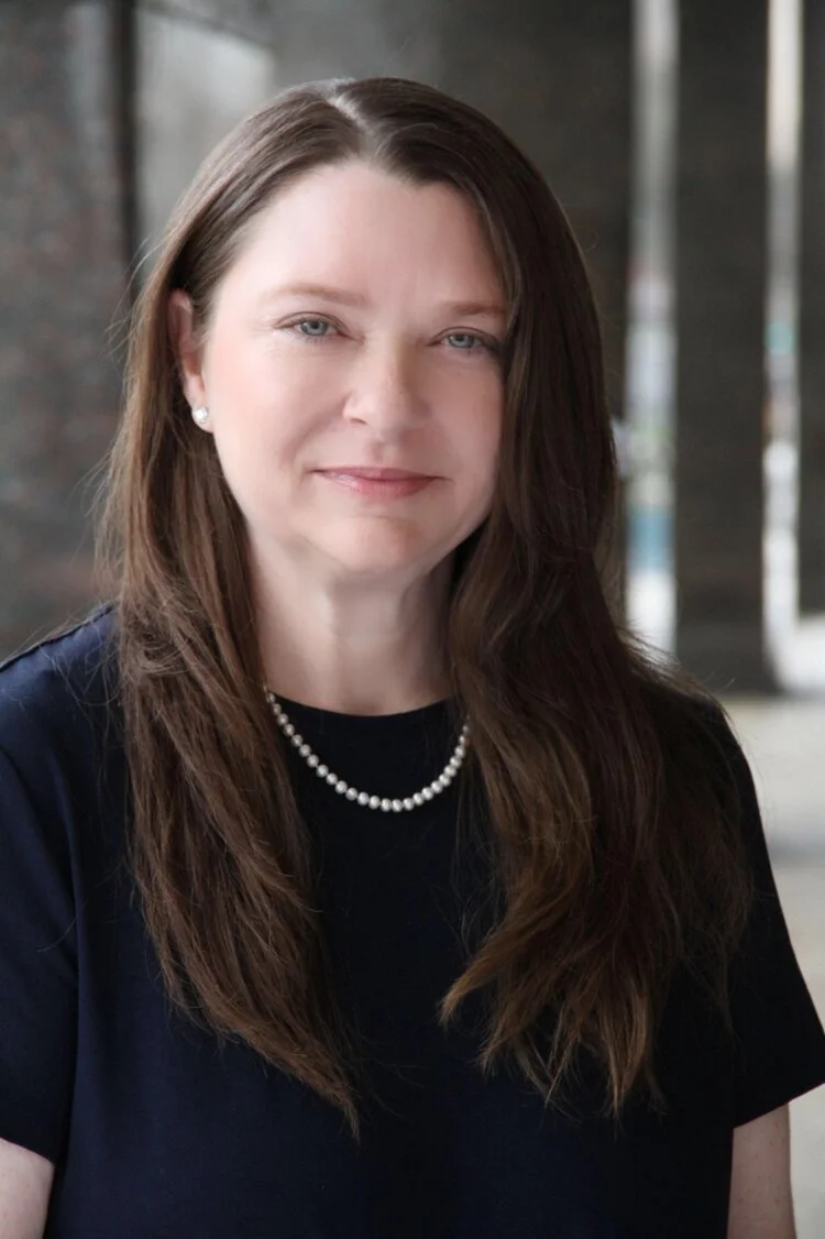 Headshot of woman with long brown hair, dark shirt, and pearls
