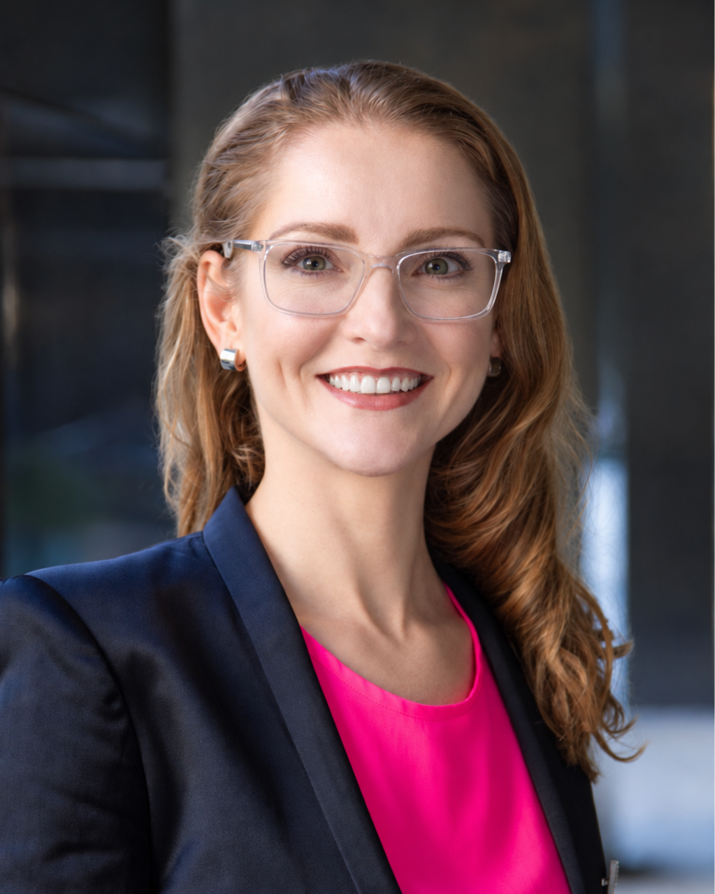 Headshot of smiling woman with long hair and glasses, wearing a dark blazer and pink shirt