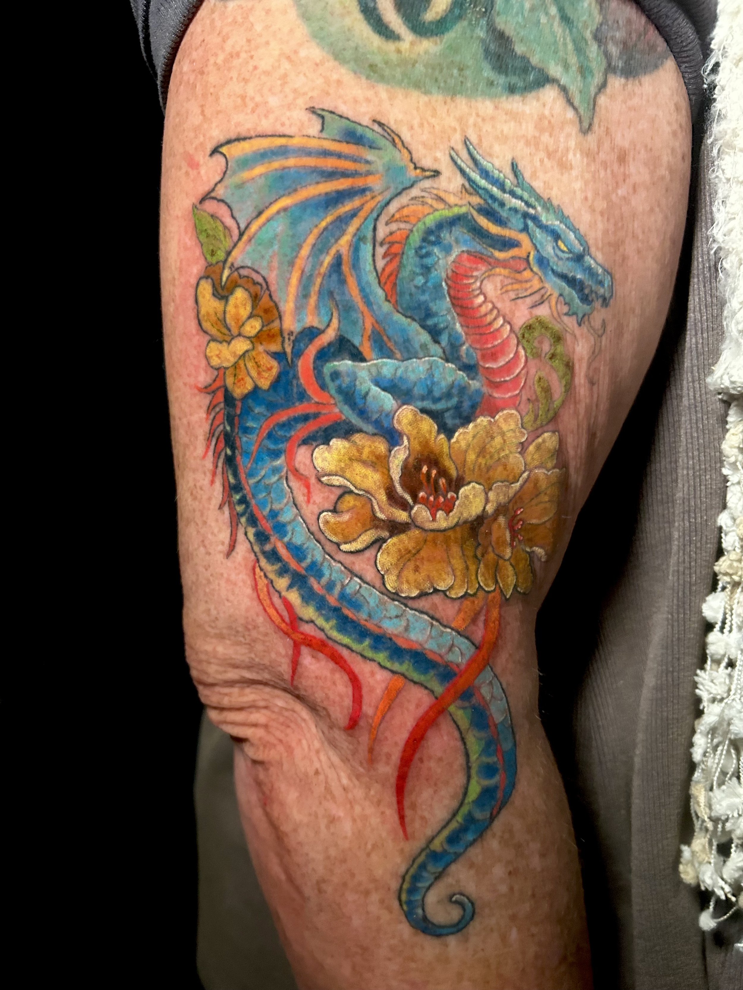Colorful tattoo of a blue dragon with flowers on skin. Tattoo by Brett Guerra.