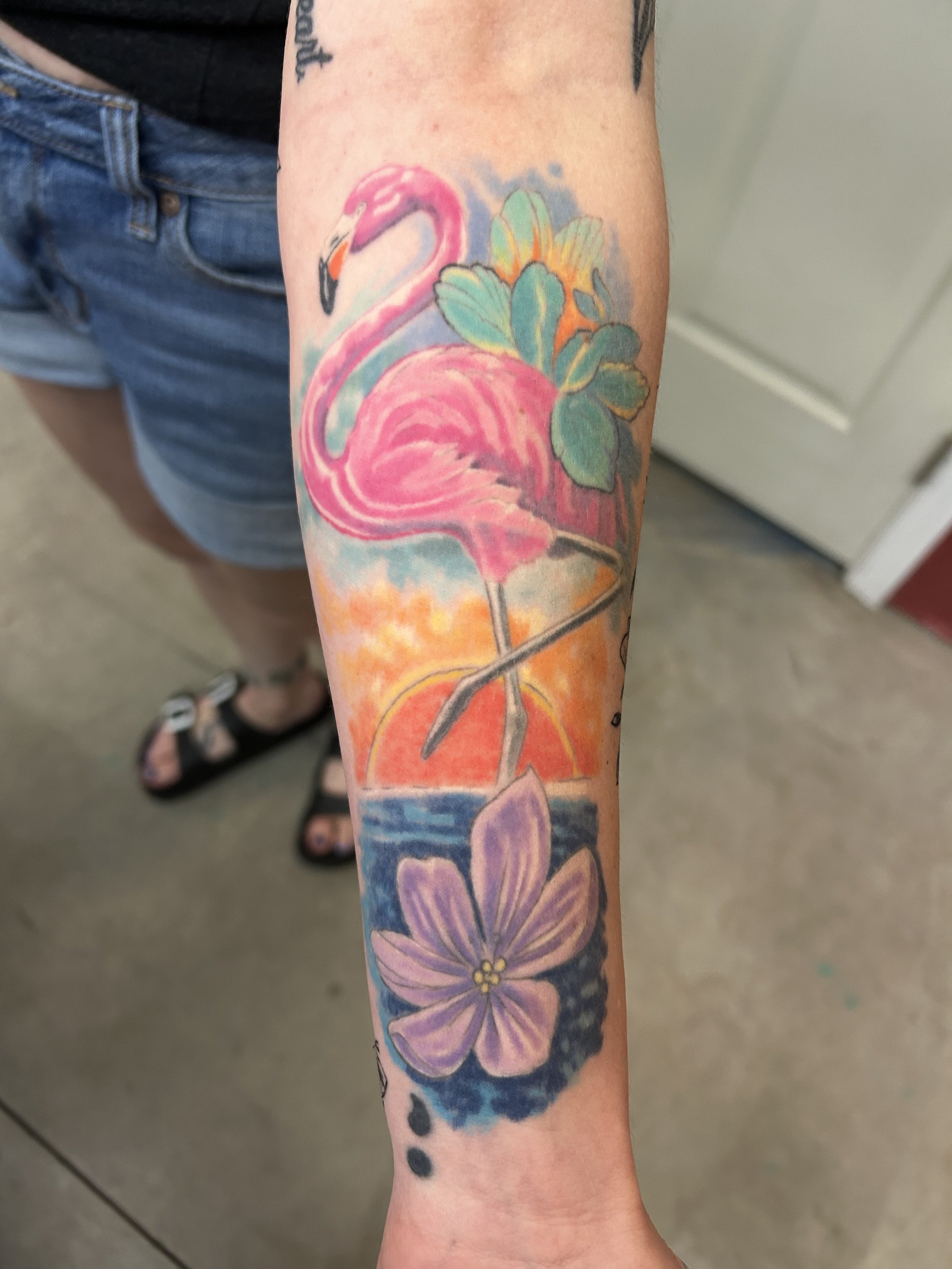 Tattoo of a pink flamingo standing in water, with a background of a sunset and tropical flowers on a person's forearm by Brett Guerra.