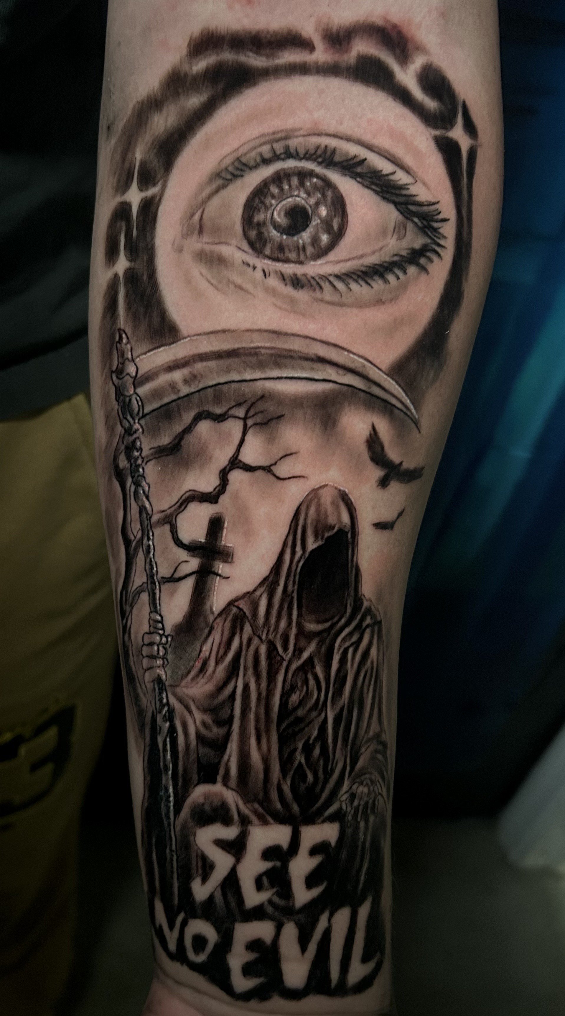 Tattoo featuring a detailed eye above a cloaked figure holding a scythe, with a cross and bare trees in the background. Text reads 'See No Evil' at the bottom. Tattoo by Brett Guerra.