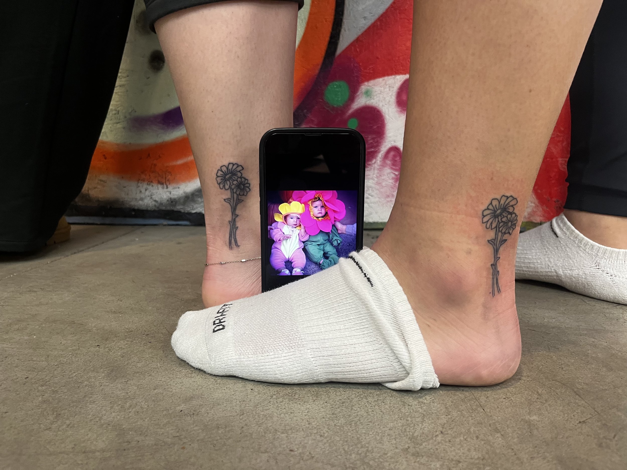 Two ankles with matching flower tattoos and a smartphone displaying a picture placed between them by Stephanie Guerra.