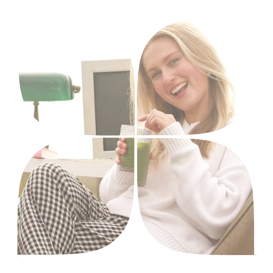 A young woman with blonde hair laughing, holding a green smoothie, sitting indoors with a computer monitor and a green desk lamp in the background.