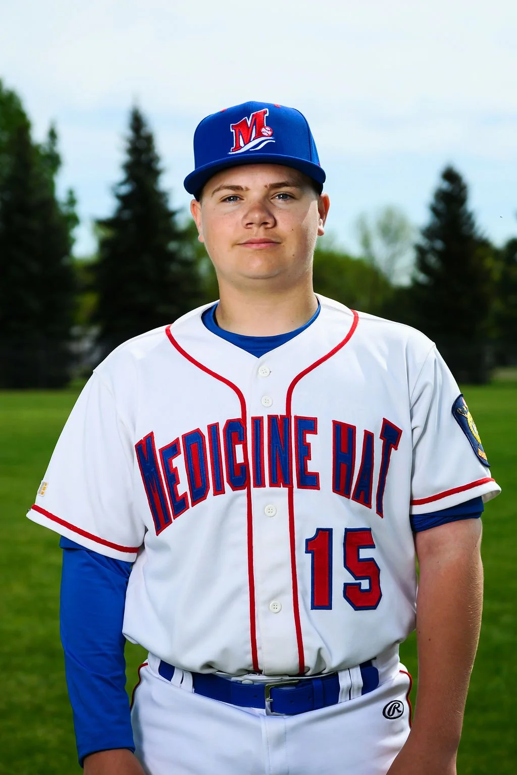 A young male baseball player in a white uniform with red and blue accents, wearing a blue cap with a red logo, standing on a baseball field with a baseball glove in hand, under a cloudy sky.