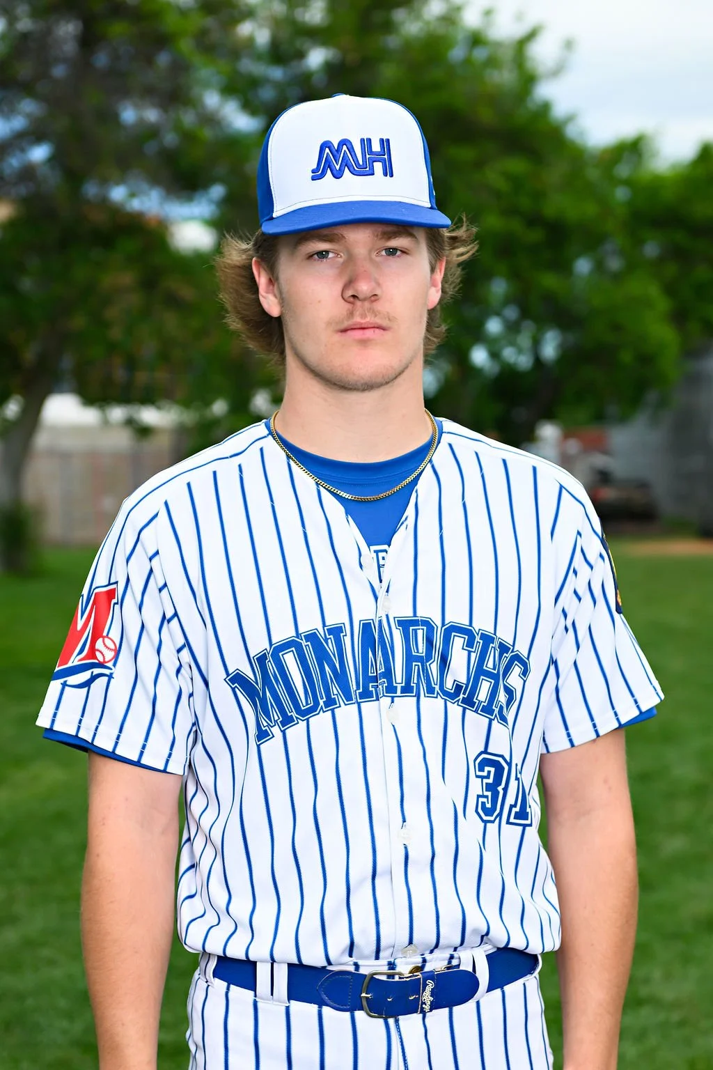 Young male baseball player standing outdoors on a baseball field, wearing a blue jersey and a blue and white baseball cap with an unreadable logo.
