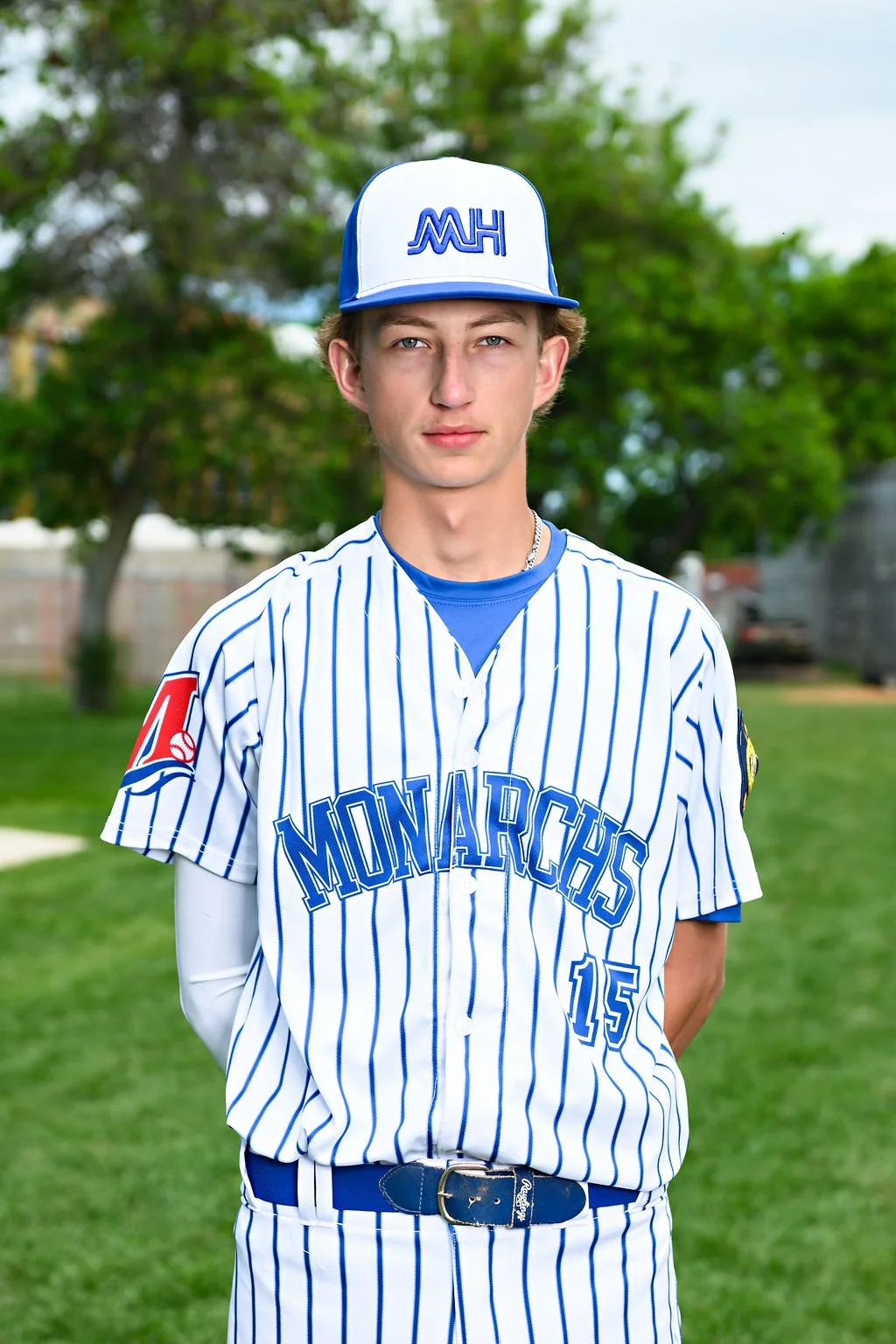 Young male baseball player in blue uniform with white accents, wearing a cap with 'MUH' logo, standing on a baseball field with trees and sky in the background.