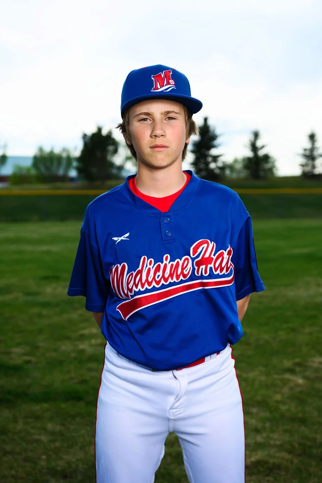 A teenage boy wearing a blue baseball cap with an 'M' logo, a blue jersey with red and white lettering that says 'Medicine Hat,' standing outdoors on a baseball field with trees and a partly cloudy sky in the background.