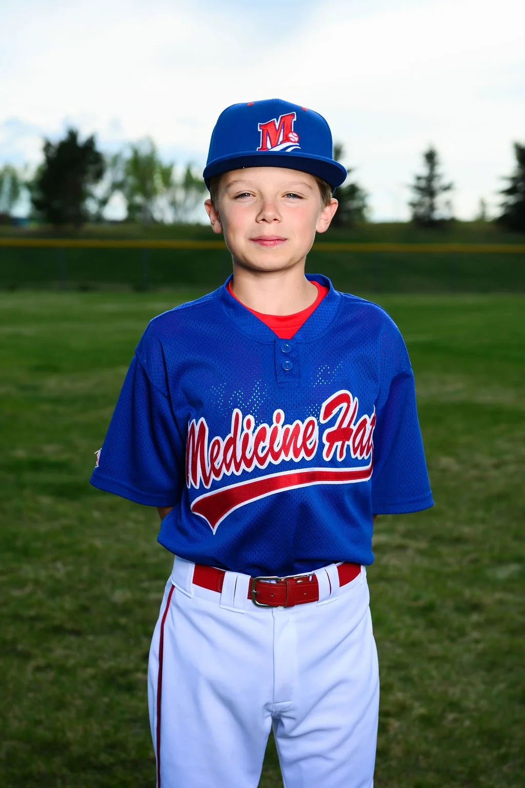 A teenage boy wearing a blue baseball jersey with 'Medicine Hat' written on it, a red shirt underneath, a navy blue cap with a red 'V' and colorful reflective sunglasses resting on the hat's brim, standing outdoors with trees and a blue sky in the background.