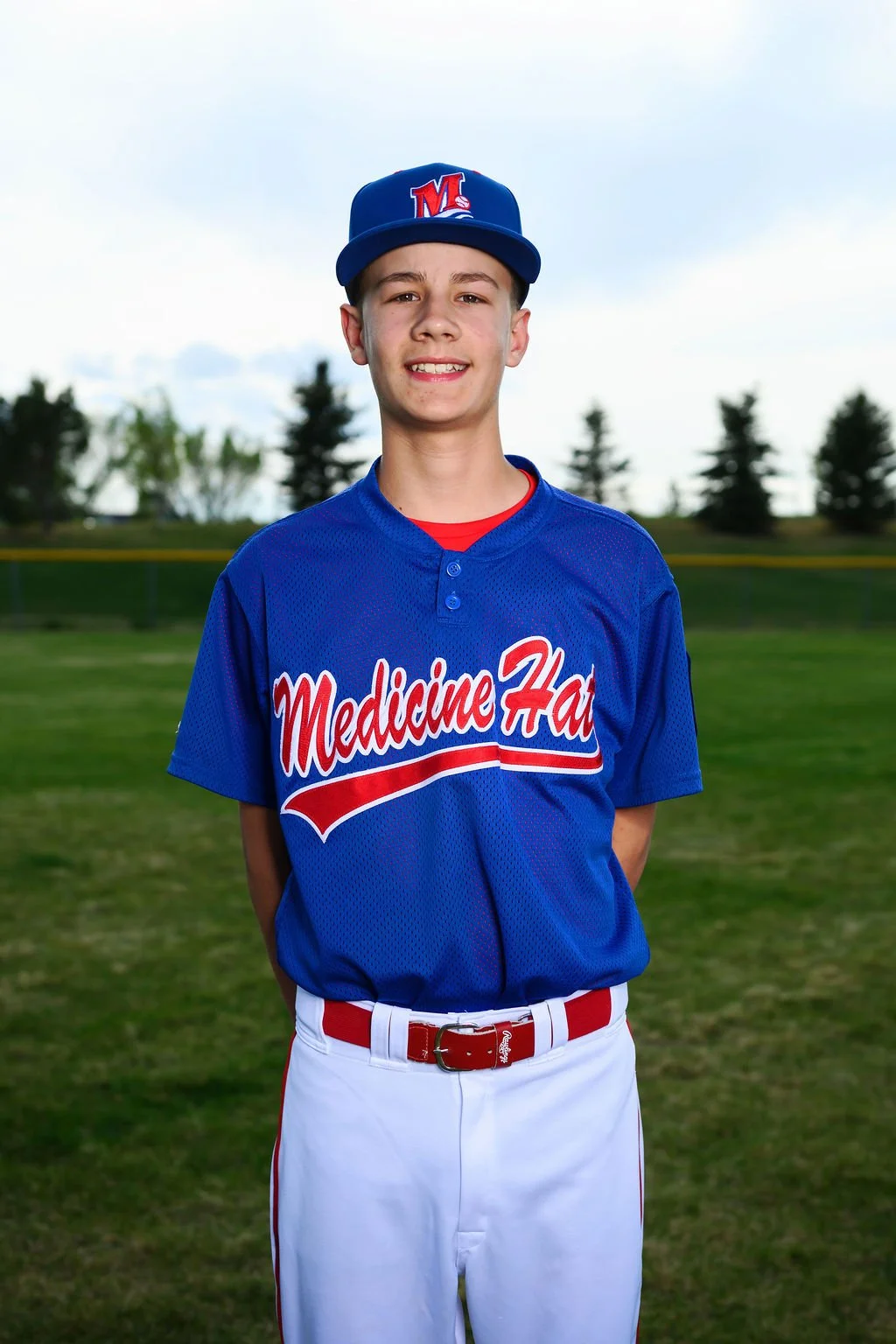 A young male baseball player standing on a field, wearing a blue jersey with red and white lettering that says 'Medicine Hat', a blue cap with a red and white 'M', and mirrored sunglasses on his cap.