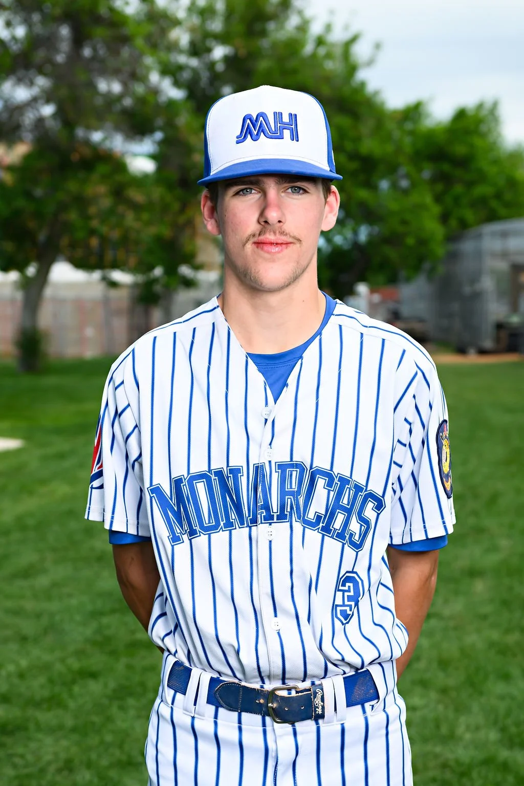 Young man in blue baseball jersey and white cap standing on a baseball field outdoors.