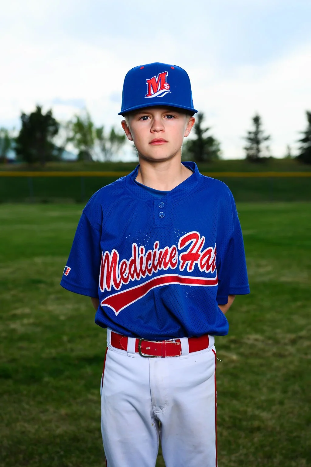 A teenage boy in a blue baseball uniform and cap standing on a baseball field with trees in the background.
