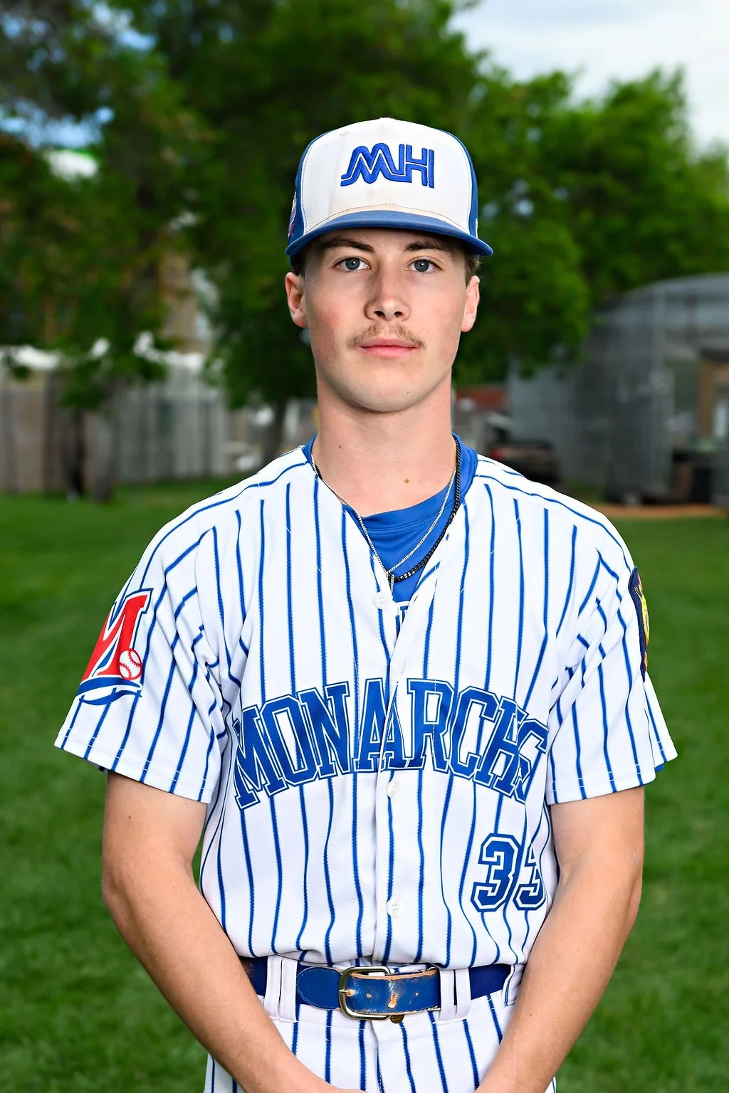 Young man standing on baseball field, wearing blue team uniform and cap with 'Mediha' logo, looking at camera.
