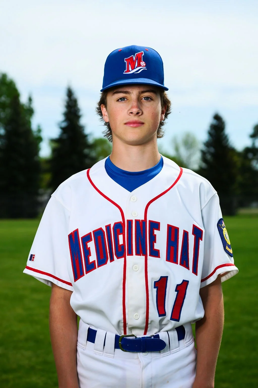 A young male baseball player wearing a white uniform with red and blue accents, a blue cap with a red and white logo, standing on a baseball field with grass and dirt, holding a glove on his left hand, looking serious.