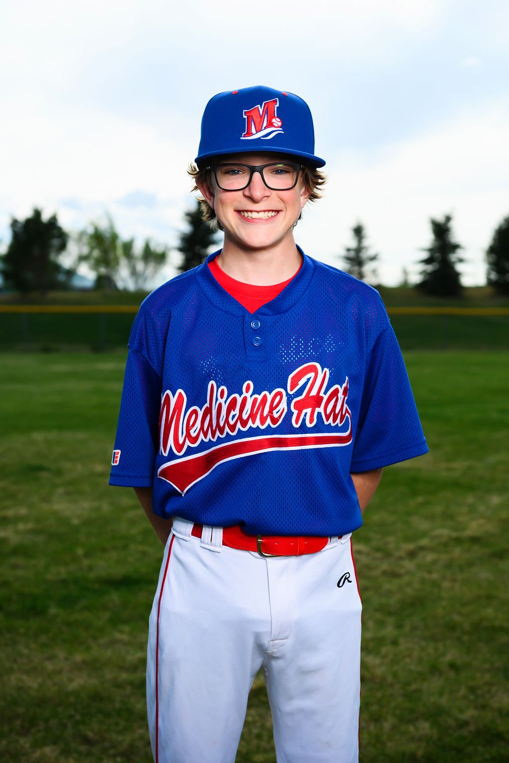 A young man wearing a blue baseball cap and jersey with 'Medicine Hat' written on it, standing outdoors on a baseball field with trees in the background.