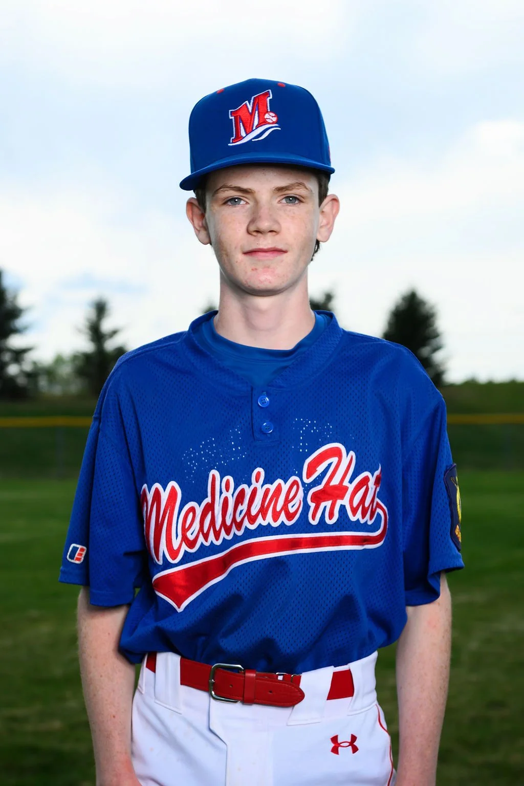 Young male baseball player in blue uniform and cap standing outdoors with trees and a partly cloudy sky in the background.