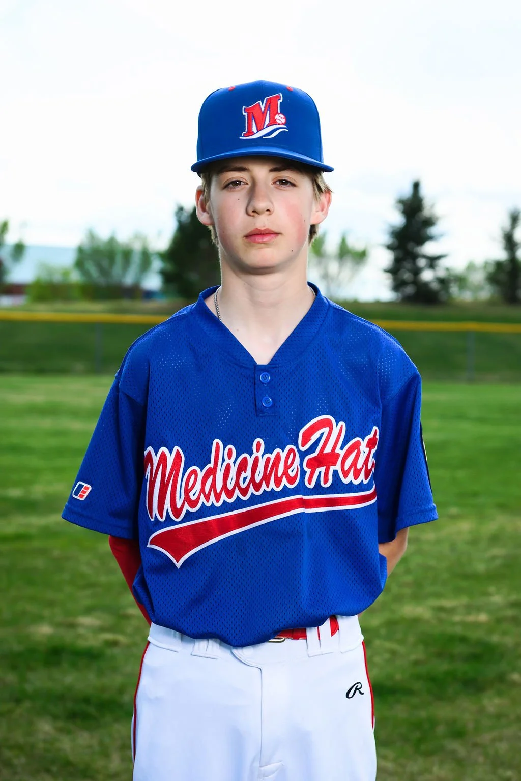A young boy wearing a blue baseball uniform and cap stands outdoors on a field with trees in the background, facing the camera.