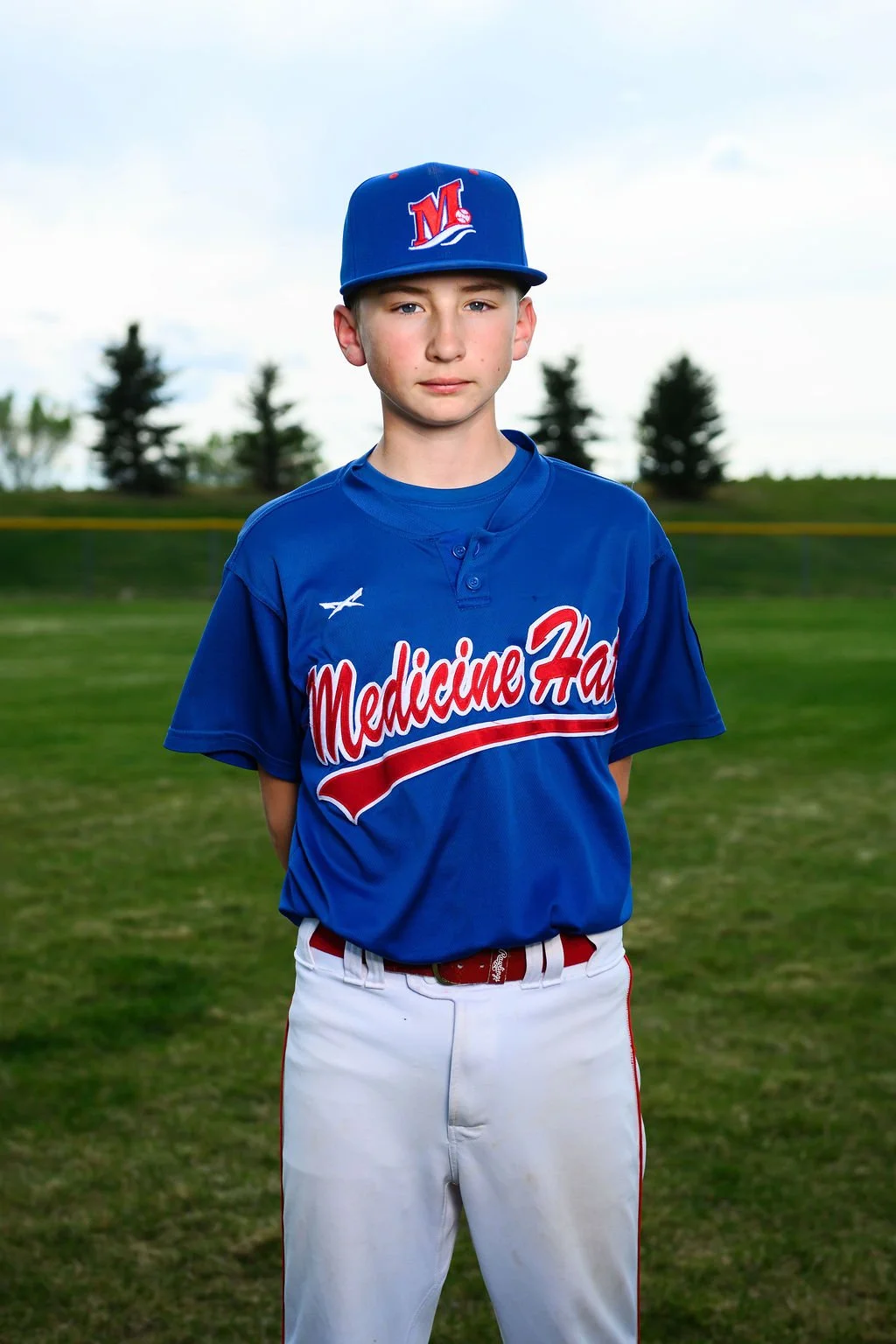 Young male baseball player wearing a blue cap with a red and white logo, blue jersey with red and white lettering, standing outdoors on a baseball field with trees in the background.