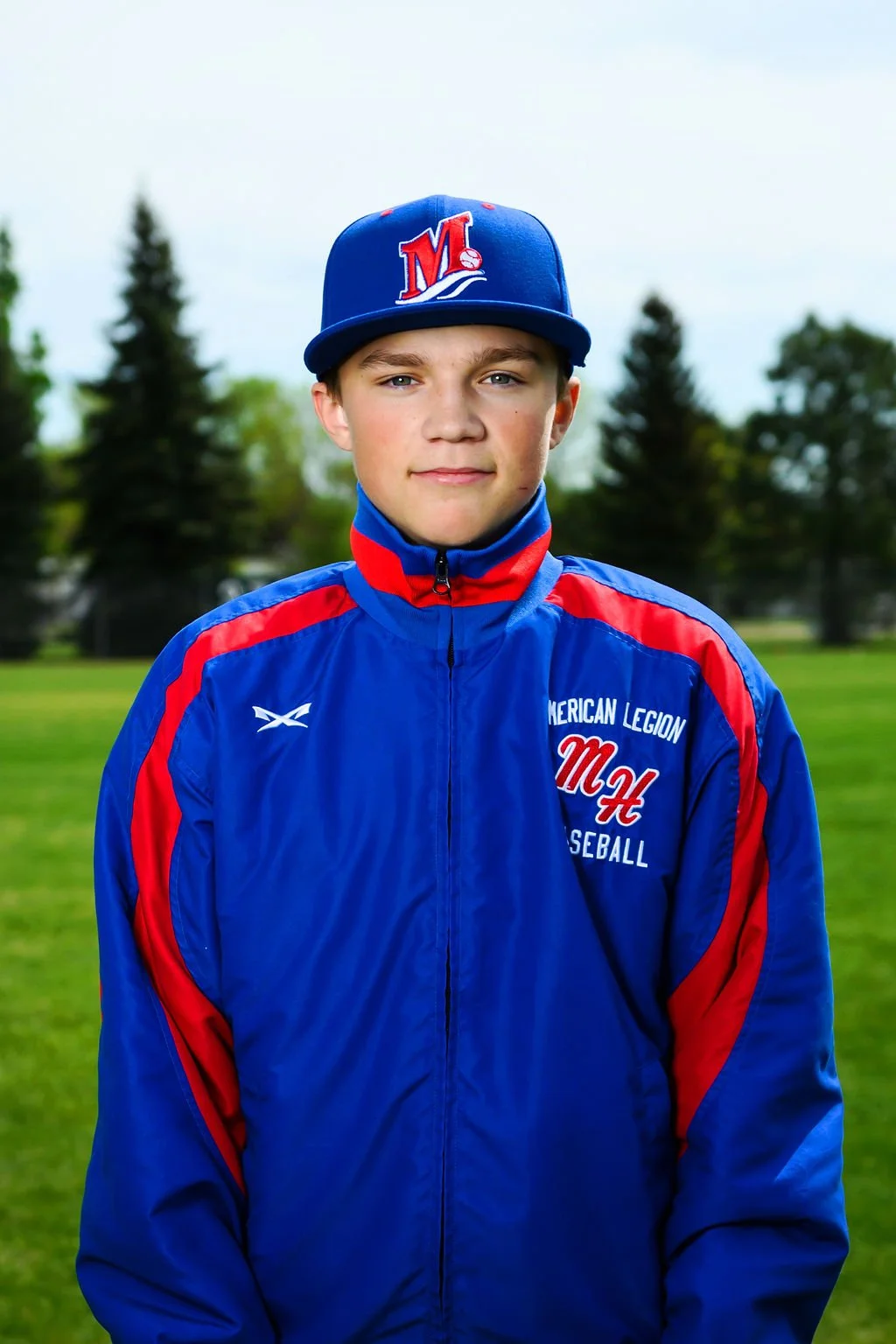 A young male baseball player in a white uniform with red and blue accents, wearing a blue cap with a red and white 'M' logo, holding a baseball glove, standing on a baseball field with trees and a cloudy sky in the background.