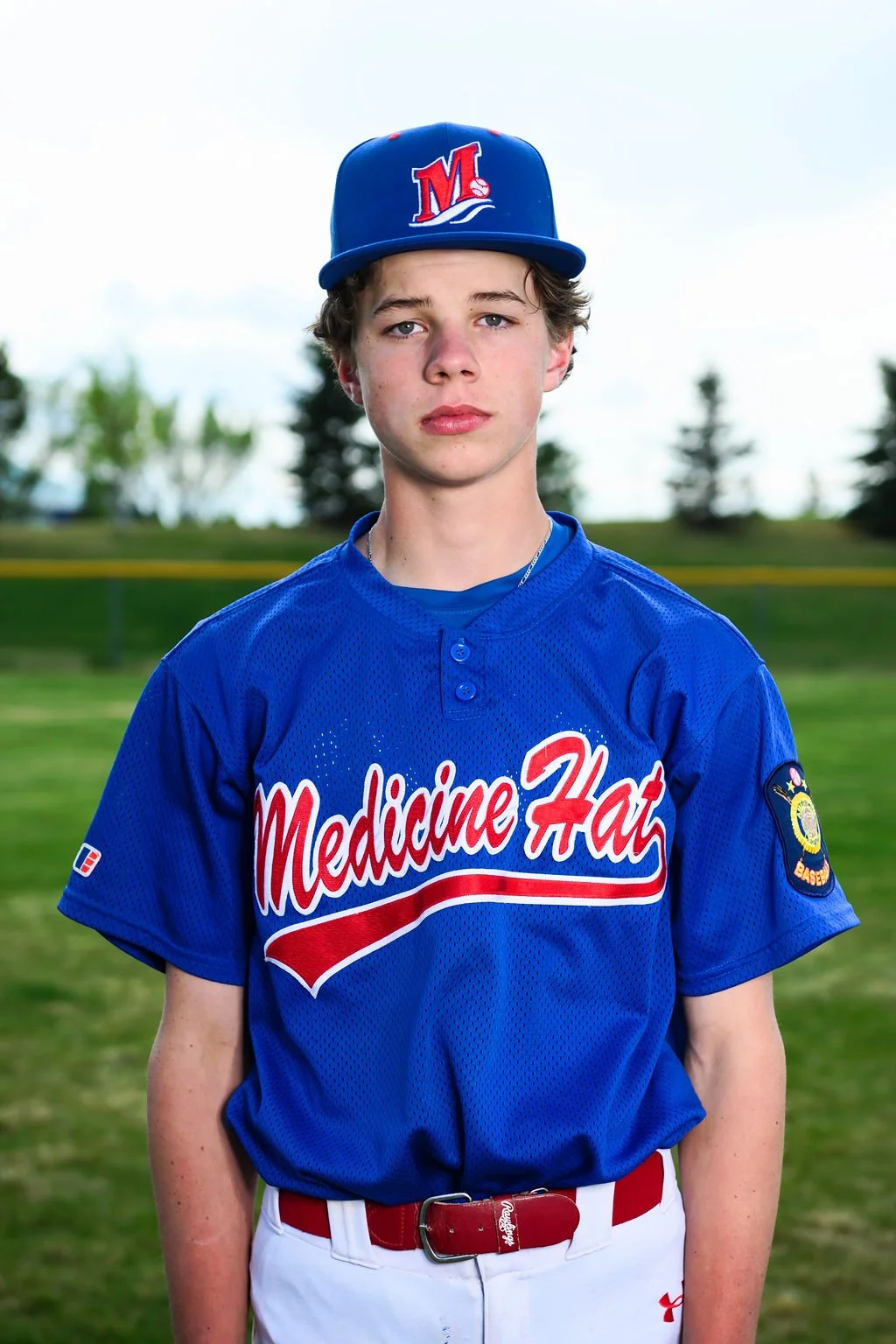 A young boy wearing a blue baseball cap with a red and white logo, and a blue sports jersey with red and white lettering that reads 'Medicine Hat', standing outdoors on a baseball field with trees in the background.