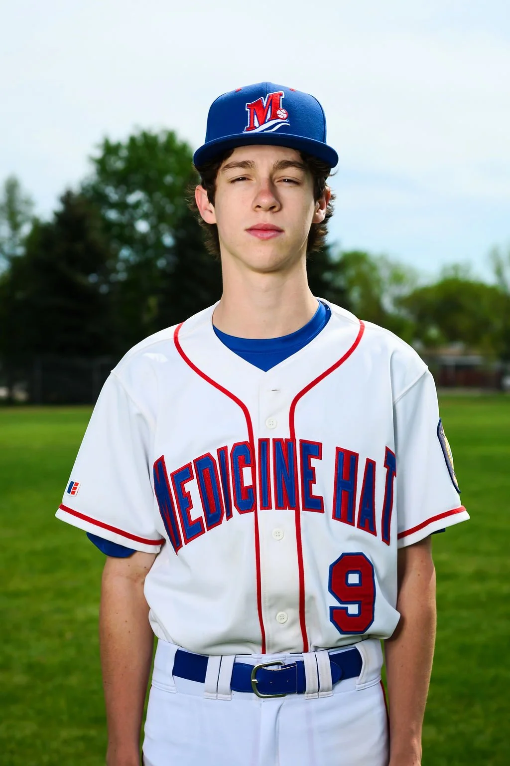 A teenage baseball player in a white uniform with red and blue accents, standing on a baseball field, holding a baseball glove, with trees and a cloudy sky in the background.